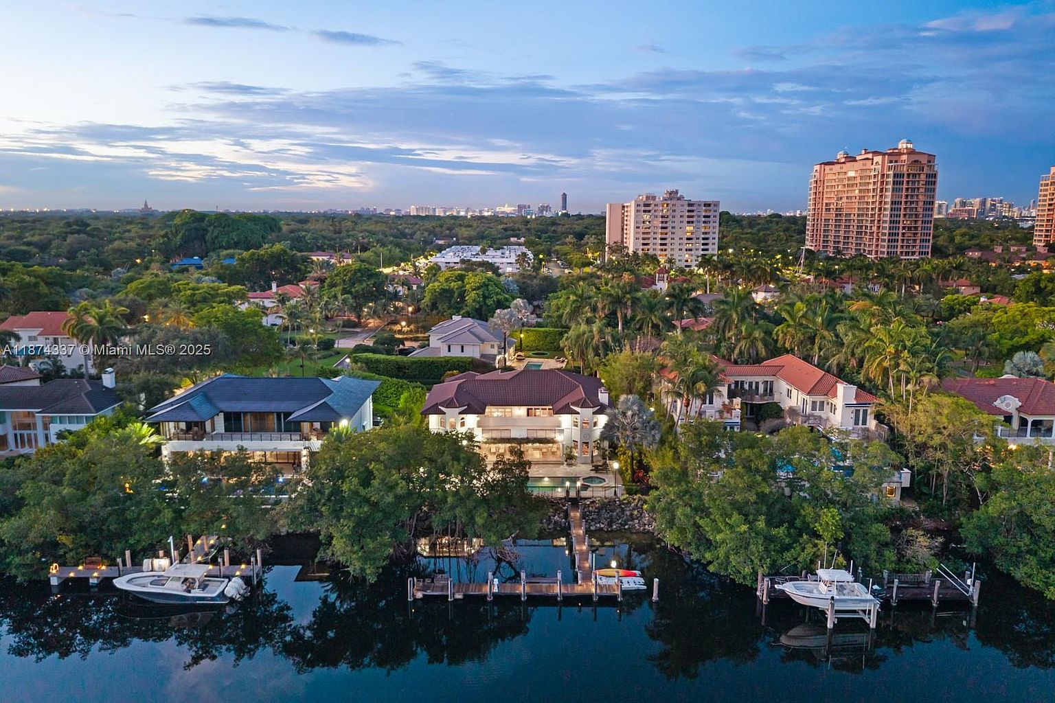 This aerial view showcases a luxurious waterfront property with a private dock and boats. The house features a well-manicured lawn, mature trees, and a swimming pool. In the background, high-rise buildings add to the upscale ambiance of the neighborhood, creating an impression of exclusivity and prestige.