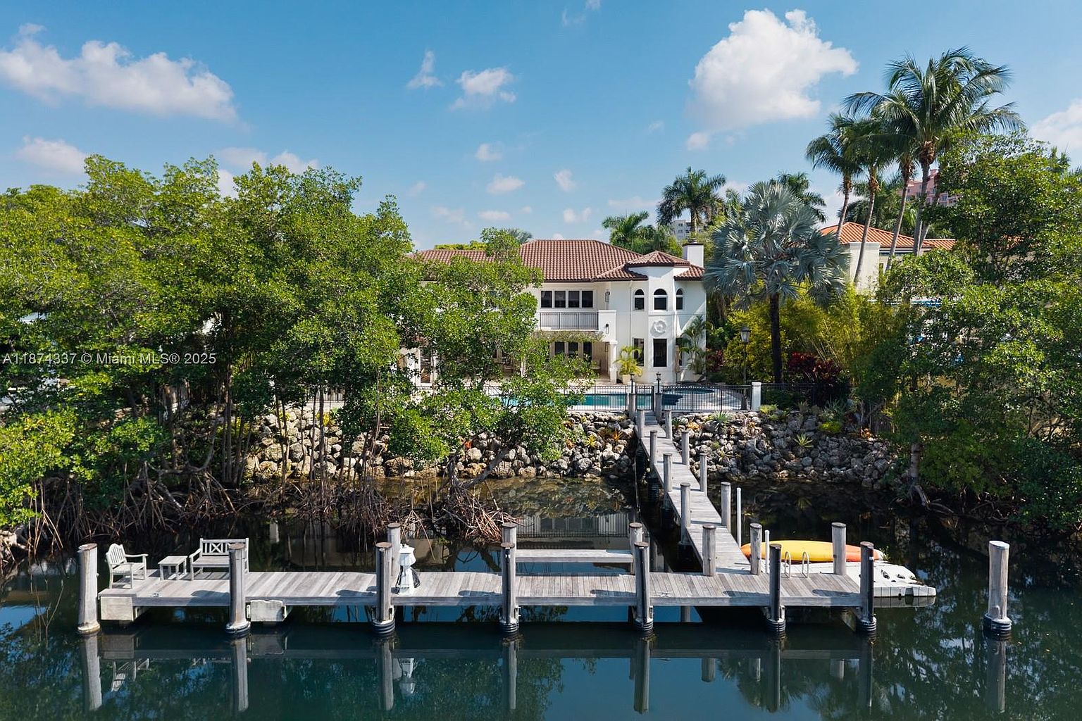 This image showcases the rear exterior of a luxurious waterfront property. The house features a Mediterranean-style design with a red tile roof, white stucco walls, and arched windows. A private dock extends into the water, offering direct access for boating and water activities. Lush landscaping surrounds the property, enhancing its privacy and appeal.