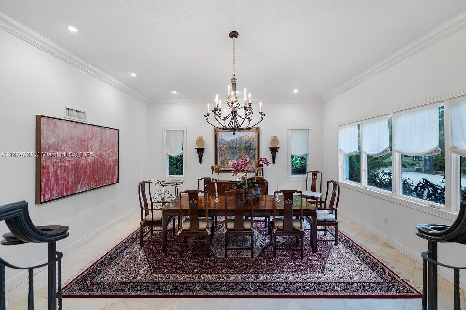This is an interior shot of a formal dining room. The room features a large wooden dining table with chairs, a chandelier, and a patterned rug. There is artwork on the walls and natural light coming in from the windows, creating a bright and elegant atmosphere.