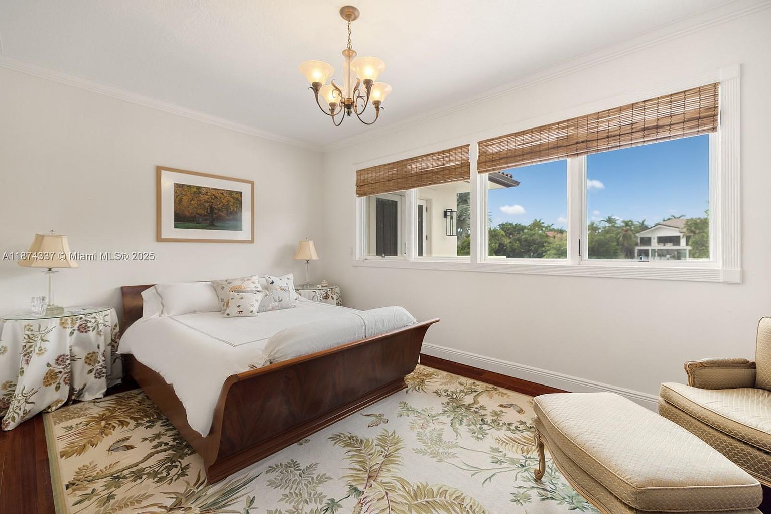 This is a well-lit primary bedroom featuring a wooden sleigh bed with white linens, complemented by a floral-patterned rug and side tables. A large window with woven shades offers a view of the exterior, while a chandelier provides overhead lighting. The room exudes a cozy and elegant atmosphere.