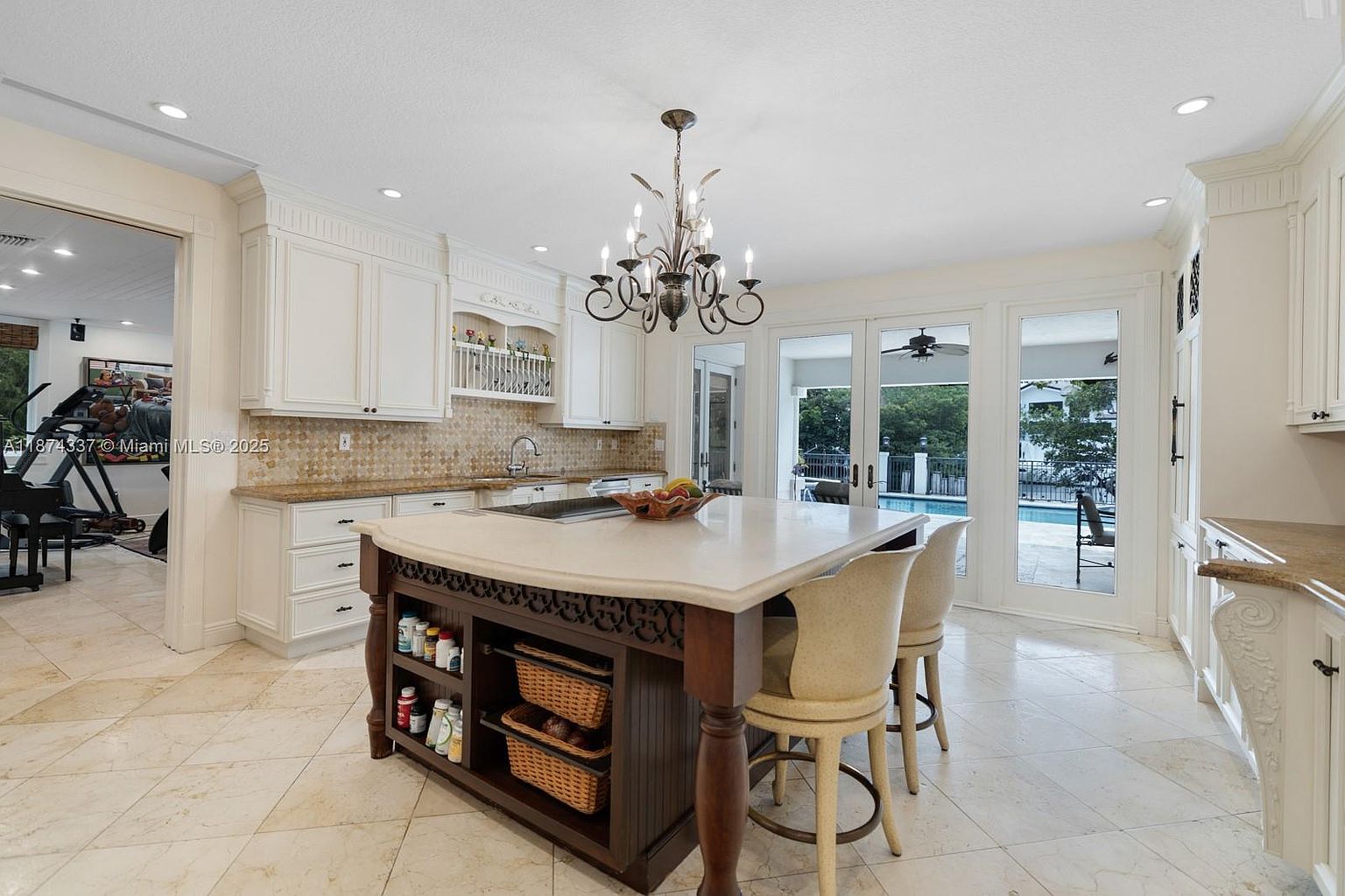 This is a bright and spacious kitchen featuring white cabinetry, a large center island with seating, and a view of the pool area through glass doors. The island has a light countertop and a dark wood base with open shelving. The flooring is light-colored tile, and a decorative chandelier hangs above the island, creating an inviting and luxurious atmosphere.
