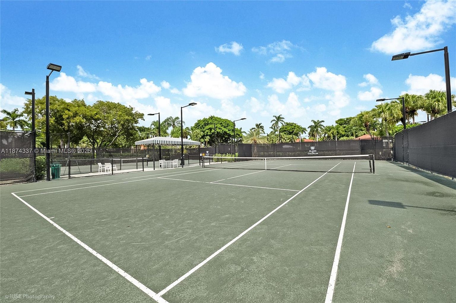 This image showcases a well-maintained tennis court, an amenity within a community. The court features a green surface with clear white lines, a net stretched across the center, and lighting fixtures for evening play. A shaded seating area is visible in the background, suggesting a comfortable space for players to relax.