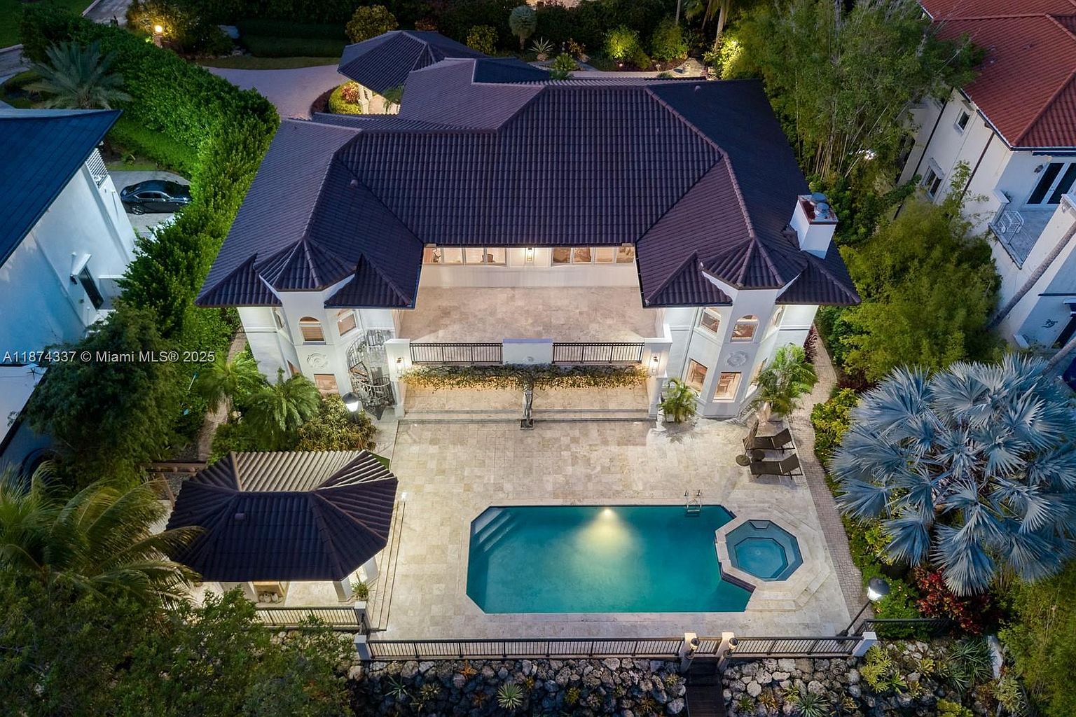This aerial view showcases a luxurious estate with a dark-tiled roof, a spacious patio, and a pristine pool and spa area. The property is surrounded by lush landscaping, providing privacy and a serene atmosphere. The architectural style is elegant and modern, emphasizing outdoor living and entertainment.