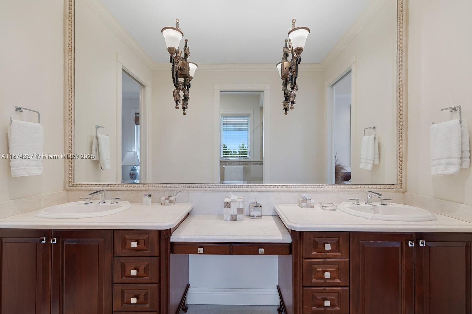 This is a primary bathroom featuring a double vanity with dark wood cabinetry and light countertops. A large, ornate mirror spans the length of the vanity, reflecting the room's elegant lighting fixtures. The overall impression is luxurious and well-appointed, suggesting a high-end property.