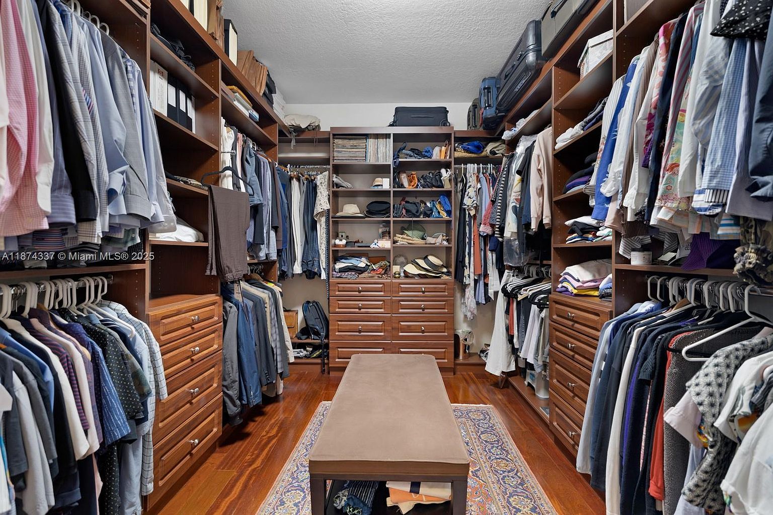 This is a well-organized walk-in closet featuring custom wooden shelving and drawers. Clothes are neatly hung and folded, showcasing ample storage space. A central ottoman provides a convenient spot for dressing, and a patterned rug adds a touch of elegance to the hardwood flooring.
