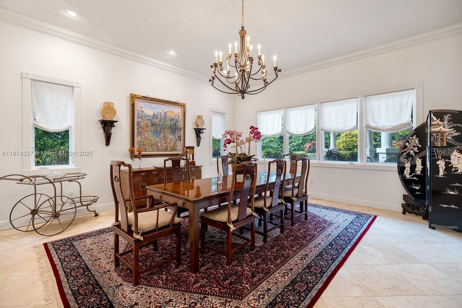 This is an interior shot of a formal dining room featuring a large wooden dining table with chairs, set on an ornate rug. A chandelier hangs above the table, and a sideboard with artwork and decorative items is positioned against the wall. The room is well-lit with natural light from the windows, creating a sophisticated and inviting atmosphere.