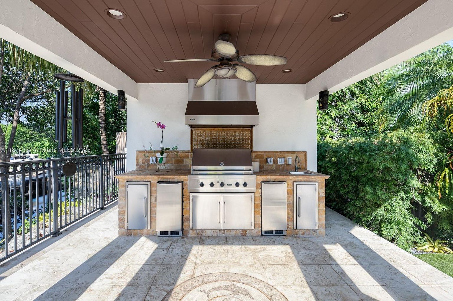 This image showcases an outdoor kitchen area on a patio or balcony. The kitchen features a built-in grill, stainless steel appliances, and stone countertops, all under a covered structure with a ceiling fan. The space is designed for outdoor entertaining and cooking, with a view of lush greenery in the background.