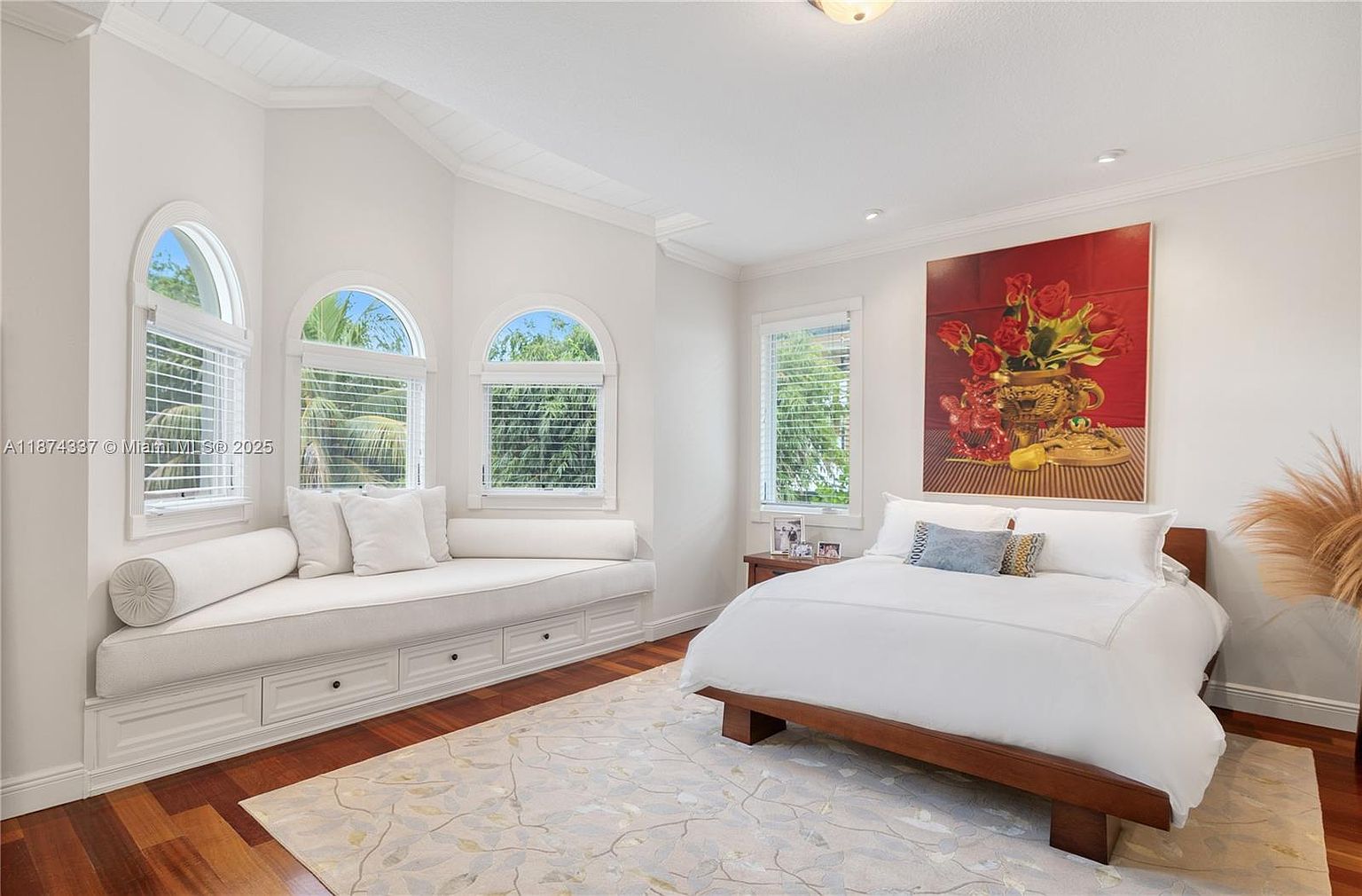 This is a bright and airy primary bedroom featuring hardwood floors, a large bed with white linens, and a seating area by the windows. A large painting adds a pop of color to the room. The overall style is elegant and comfortable, creating a relaxing atmosphere.