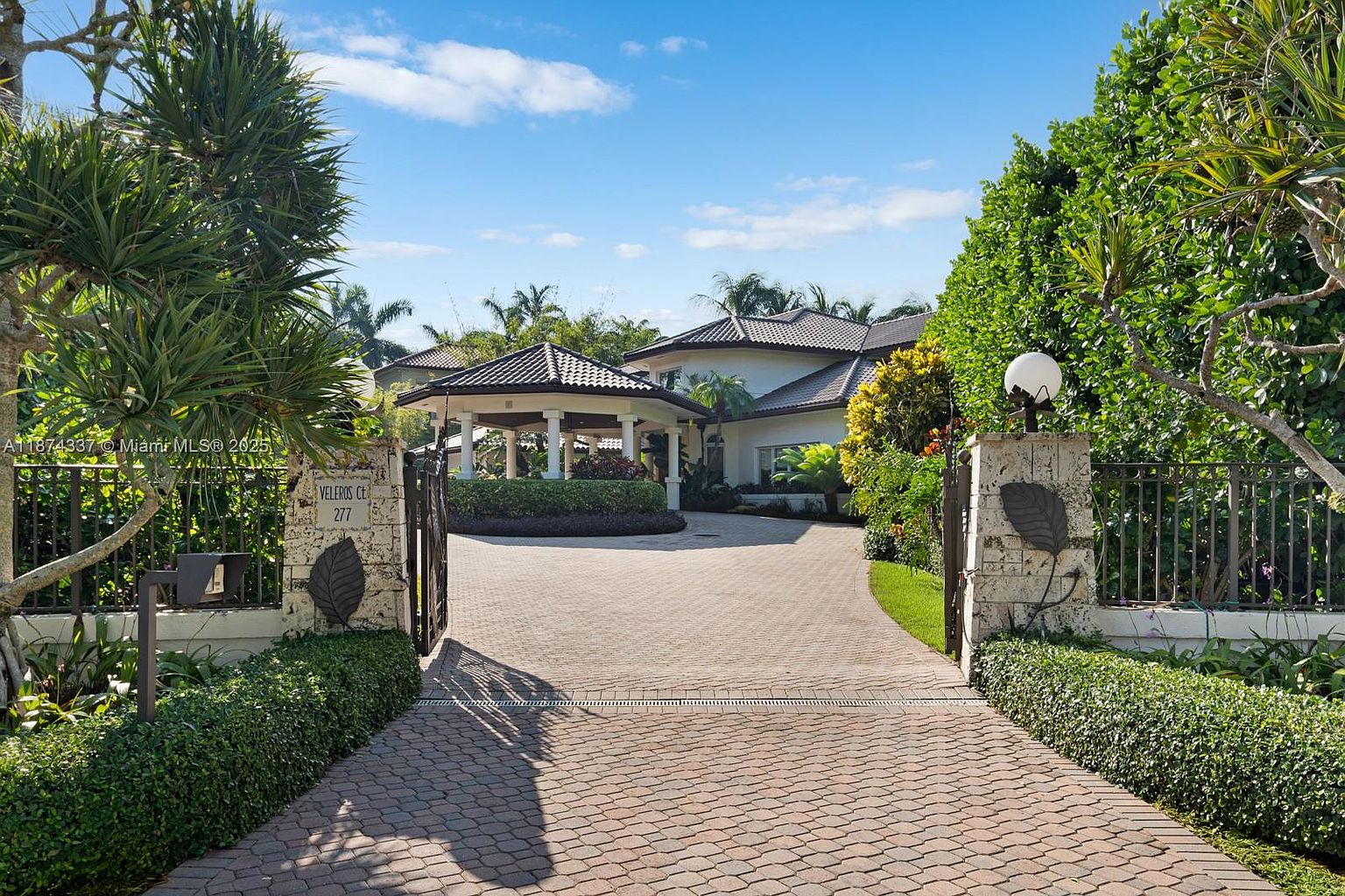 The image showcases the grand entryway of a luxurious property. A brick driveway leads through an ornate gate flanked by stone pillars and lush greenery. In the background, a covered portico and the main house are visible, suggesting a spacious and well-maintained estate. The perspective is from the entrance looking towards the house.