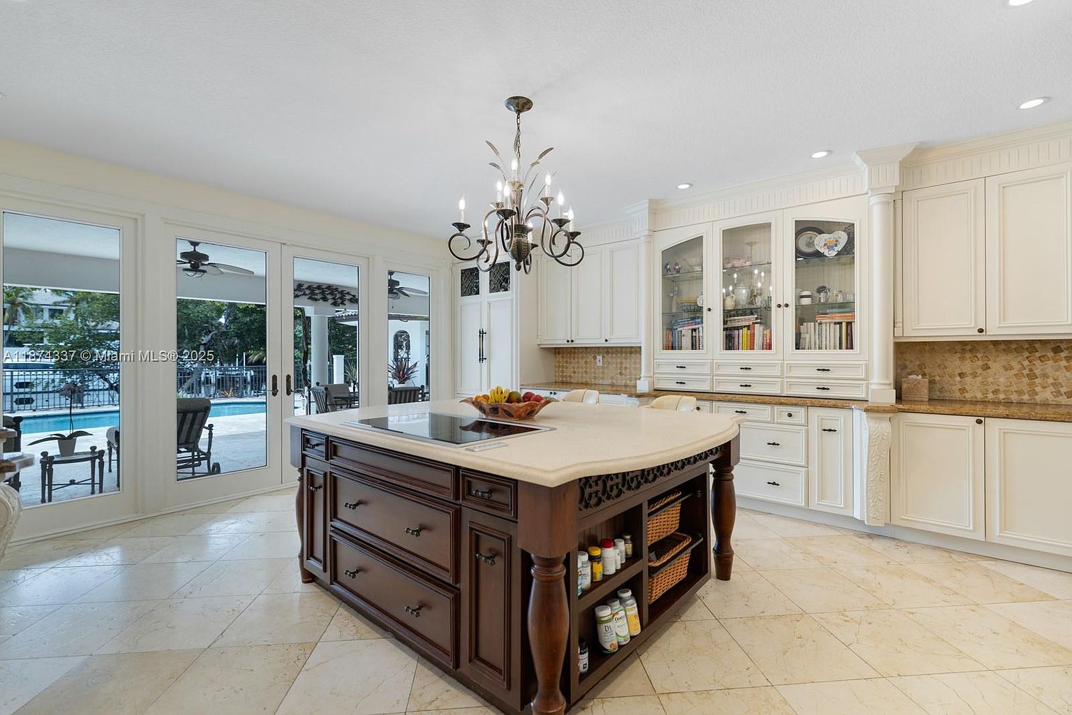 This is a well-lit kitchen featuring a large dark wood island with a light countertop and built-in cooktop. The kitchen also includes white cabinetry, glass-front display cabinets, and a decorative chandelier. Large glass doors offer a view of the outdoor pool area, creating a bright and inviting space.
