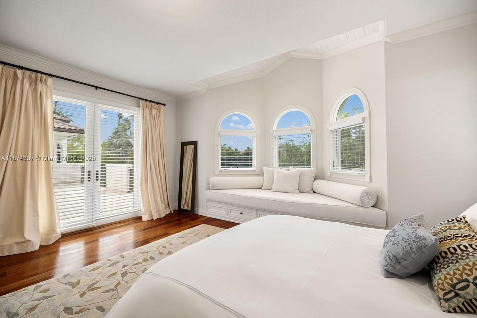 This is a bright and airy primary bedroom featuring a large bed with white linens and decorative pillows. The room has hardwood floors, a neutral-toned rug, and a built-in window seat beneath arched windows. Sliding glass doors lead to a balcony, and the overall style is elegant and comfortable.