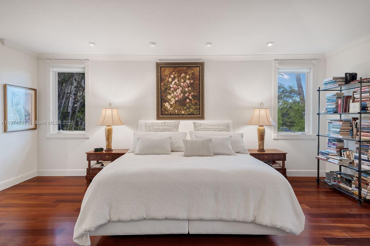 This is a well-lit primary bedroom featuring a large bed with white linens, flanked by matching nightstands and lamps. A floral painting hangs above the bed, and windows on either side of the bed provide natural light. A bookshelf filled with books is visible on the right side of the room, adding a touch of character.
