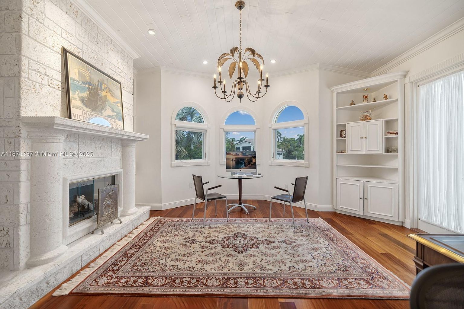 This interior shot showcases a cozy living room with a prominent white brick fireplace and a decorative mantle. The room features hardwood floors covered by a large, ornate rug, and a chandelier hangs from the white paneled ceiling. Arched windows provide natural light, and built-in shelving adds to the room's character, creating a warm and inviting atmosphere.