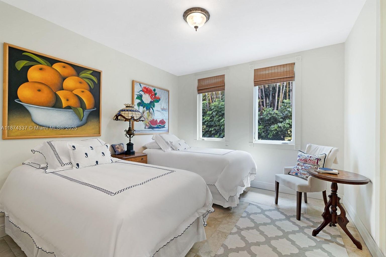 This is a bright and inviting guest bedroom featuring two twin beds with white linens and nautical-themed accents. Artwork adorns the walls, and natural light streams in through the windows, creating a serene atmosphere. A small table and chair provide a cozy seating area, enhancing the room's functionality and charm.