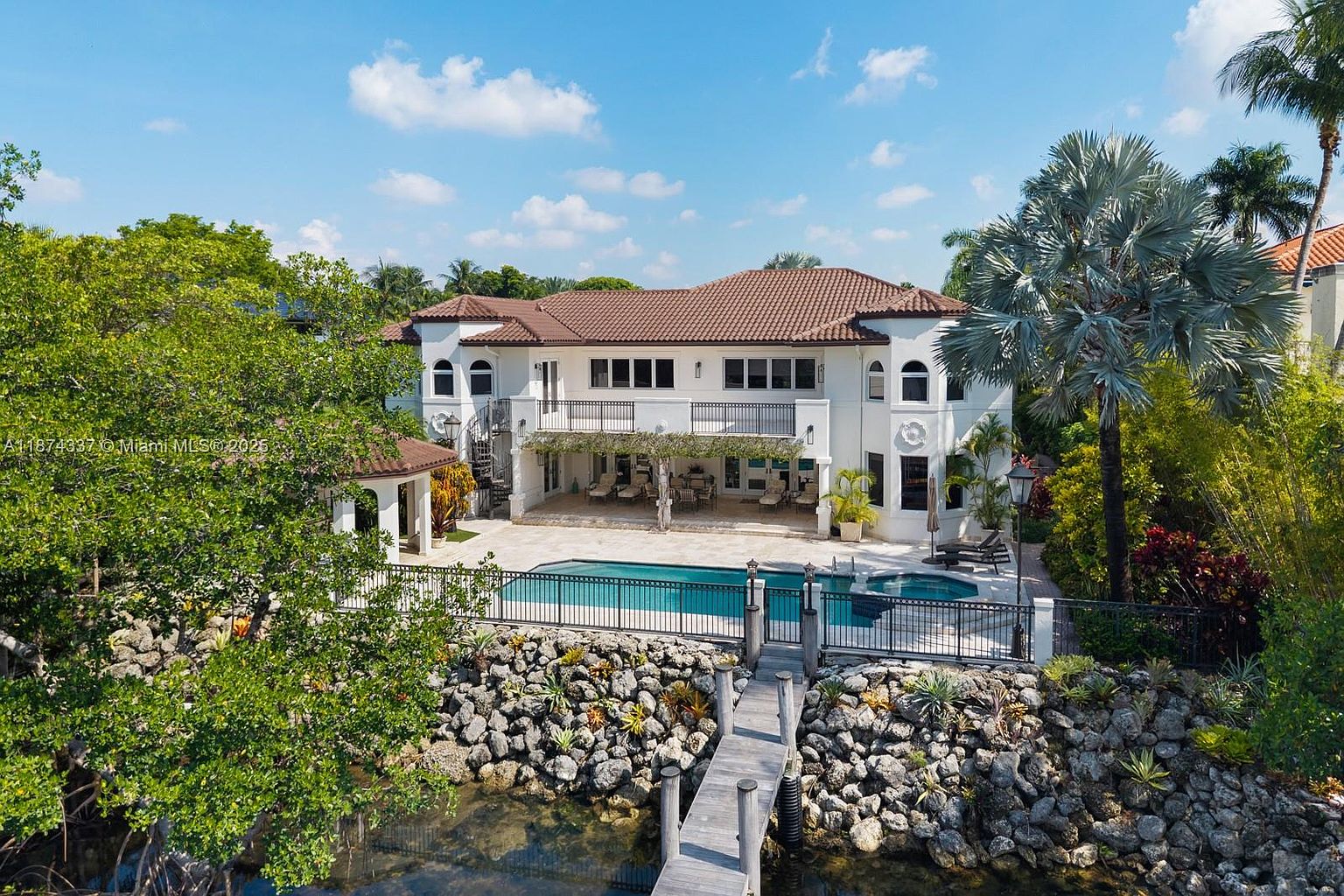 This image showcases the rear exterior of a luxurious two-story home with a Mediterranean-inspired design, featuring a terracotta tile roof and white stucco walls. A spacious patio area overlooks a swimming pool and a waterfront with a private dock, creating an inviting outdoor living space. Lush landscaping surrounds the property, enhancing its privacy and appeal.