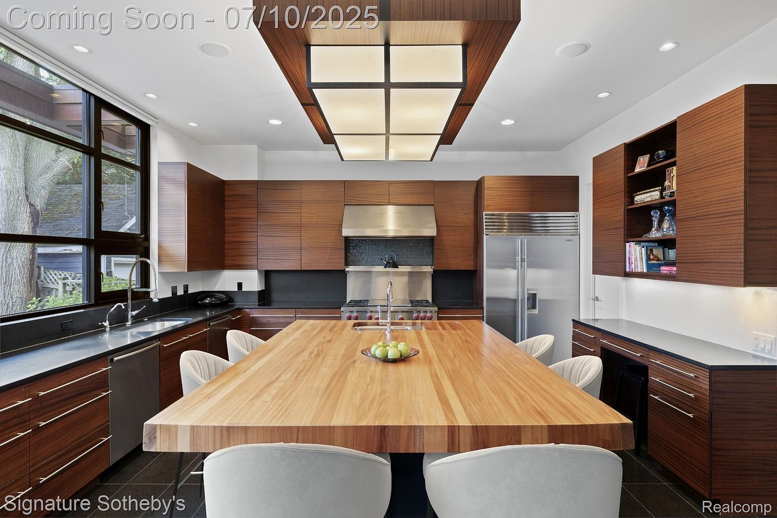 This is a modern kitchen featuring sleek, dark wood cabinetry and a large, light wood island with seating. Stainless steel appliances, including a large refrigerator and range hood, add a contemporary touch. The room is well-lit with recessed lighting and a unique overhead fixture, creating a bright and inviting space.