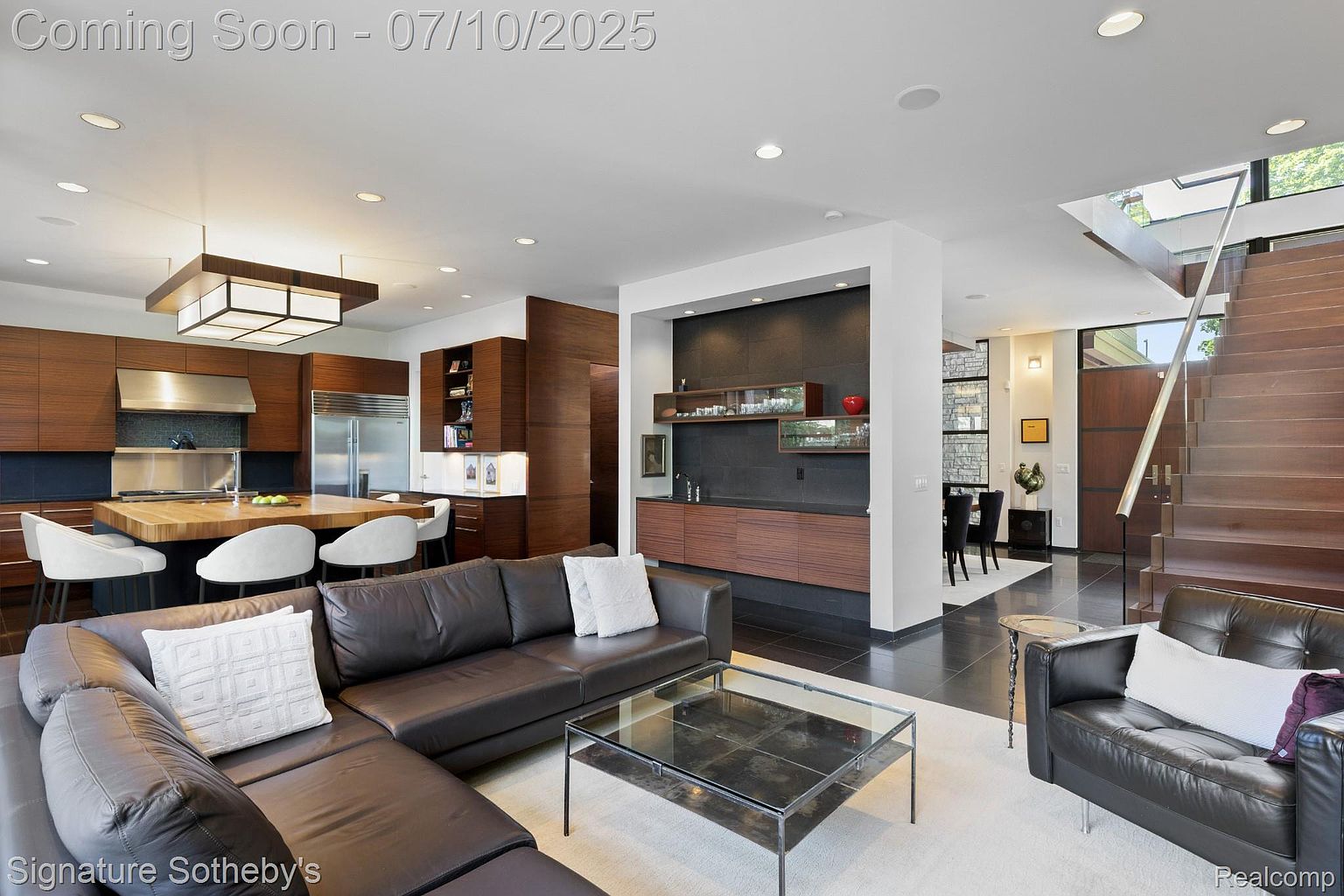 This is an interior shot of a modern living room that seamlessly blends into the kitchen and dining area. The living room features a large, dark leather sectional sofa, a glass coffee table, and a black leather armchair, all set on a light-colored rug. The kitchen boasts wooden cabinetry and a central island with bar seating, while the dining area is visible in the background, creating an open and inviting space.
