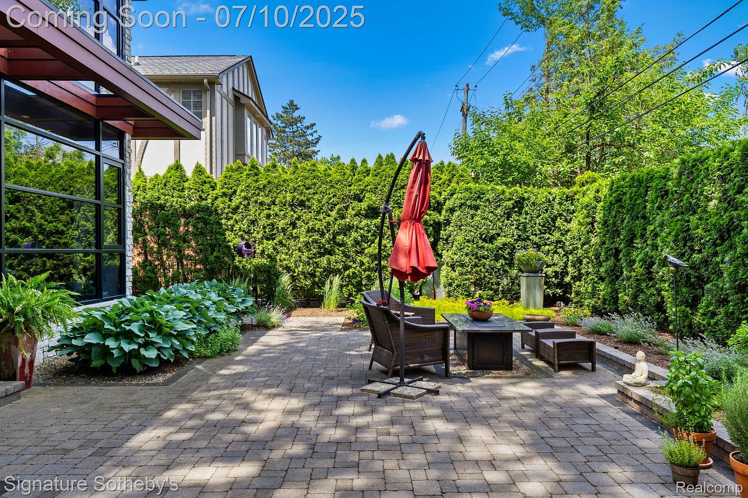 This image showcases a charming outdoor patio area, featuring a brick-paved surface, comfortable wicker seating, and a red umbrella for shade. Lush green hedges provide privacy and create a serene backyard setting. The patio is decorated with potted plants and a small Buddha statue, adding to the tranquil ambiance.