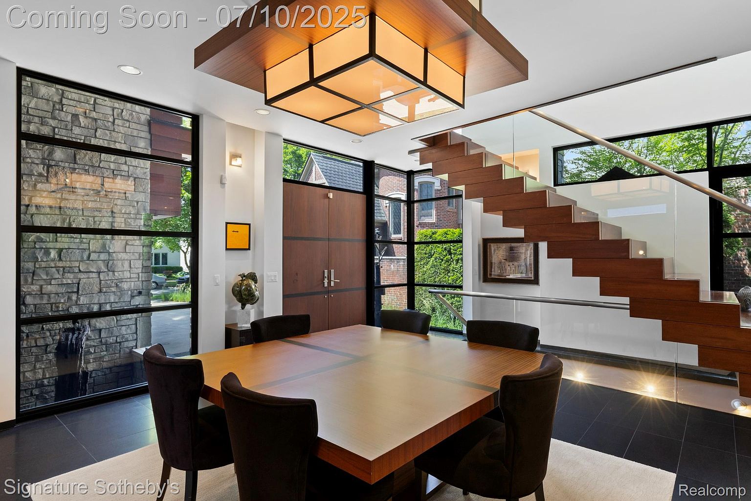 This interior shot showcases a modern dining room with a large square wooden table surrounded by six dark upholstered chairs. The room features striking architectural elements, including a floating staircase with glass railings and large windows that provide ample natural light and views of the exterior. A unique ceiling light fixture adds a touch of contemporary elegance.