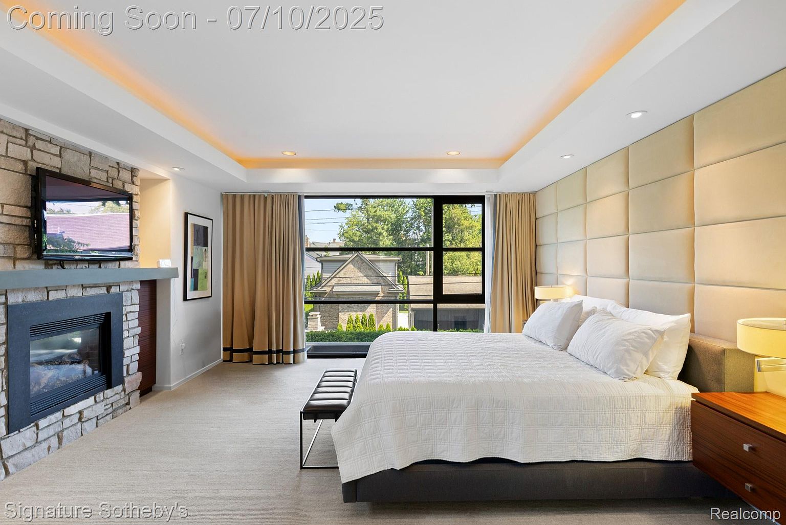 This is a luxurious primary bedroom featuring a king-size bed with white linens, a padded headboard wall, and a modern fireplace with a mounted television. Large windows offer natural light and views of the outdoors, complemented by elegant curtains. The room exudes a sense of comfort and sophistication, perfect for relaxation.