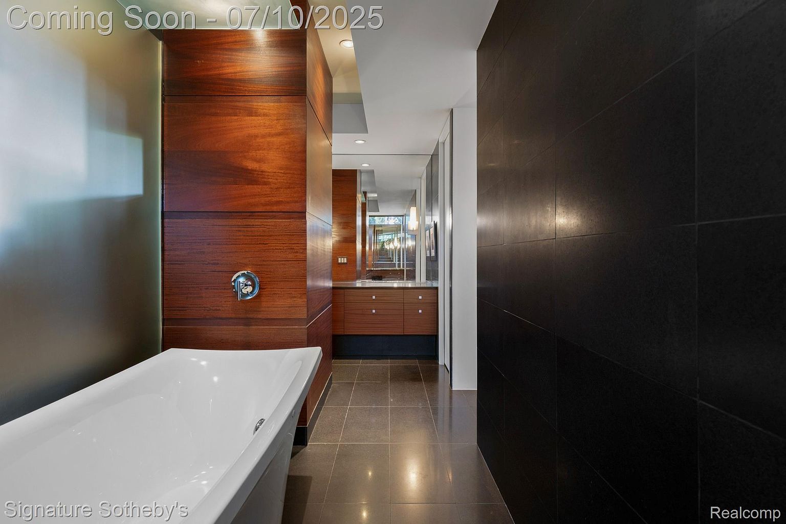 This is a modern primary bathroom featuring a freestanding white bathtub, dark wood cabinetry, and dark tiled walls. The space is well-lit and appears luxurious, with a clean and minimalist design. The perspective gives a sense of depth and spaciousness.