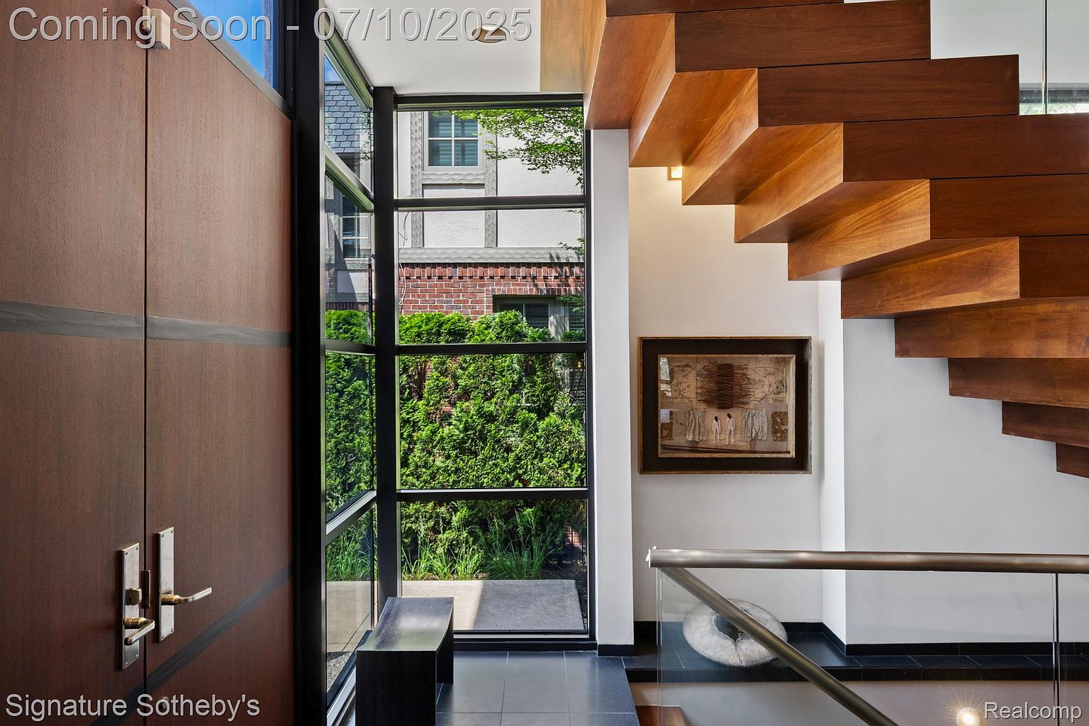 This interior shot showcases a modern hallway and staircase. The floating wooden stairs are a striking architectural feature, complemented by a glass railing. A large window provides natural light and a view of the exterior greenery, while a dark wood door adds a touch of elegance.