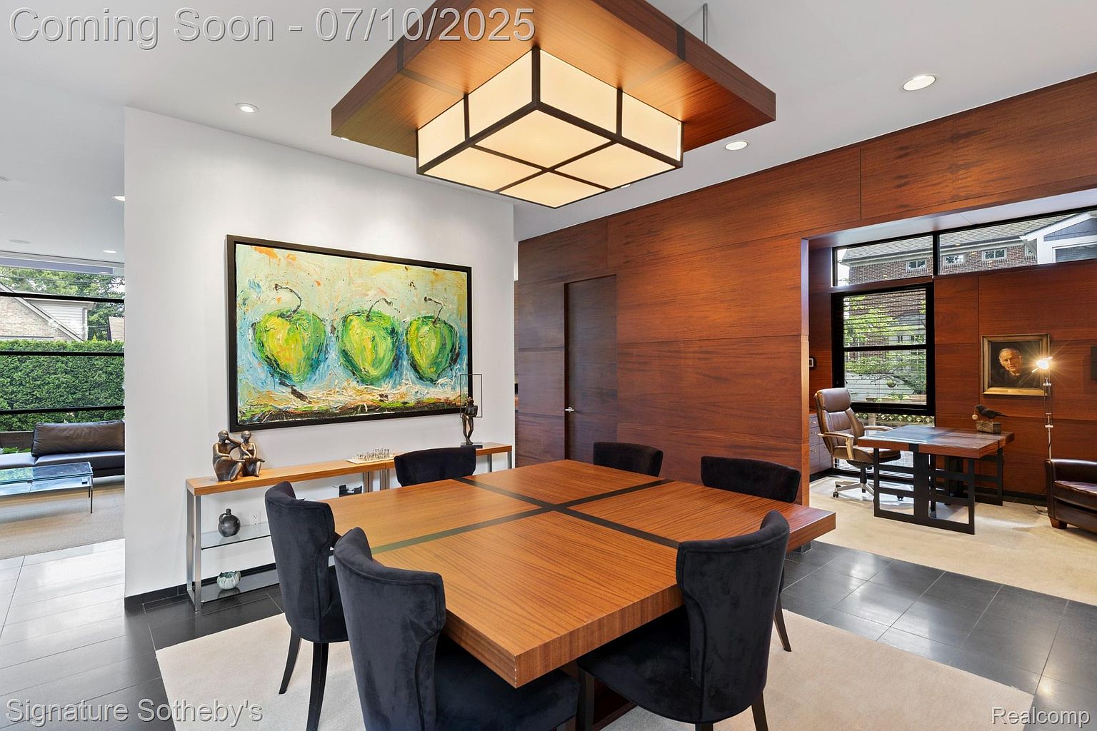 This interior shot showcases a modern dining room with a large square wooden table surrounded by plush black chairs. A striking piece of art hangs above a console table, and a unique square light fixture illuminates the space. The room seamlessly transitions into an office area with wood paneling, creating a sophisticated and functional atmosphere.