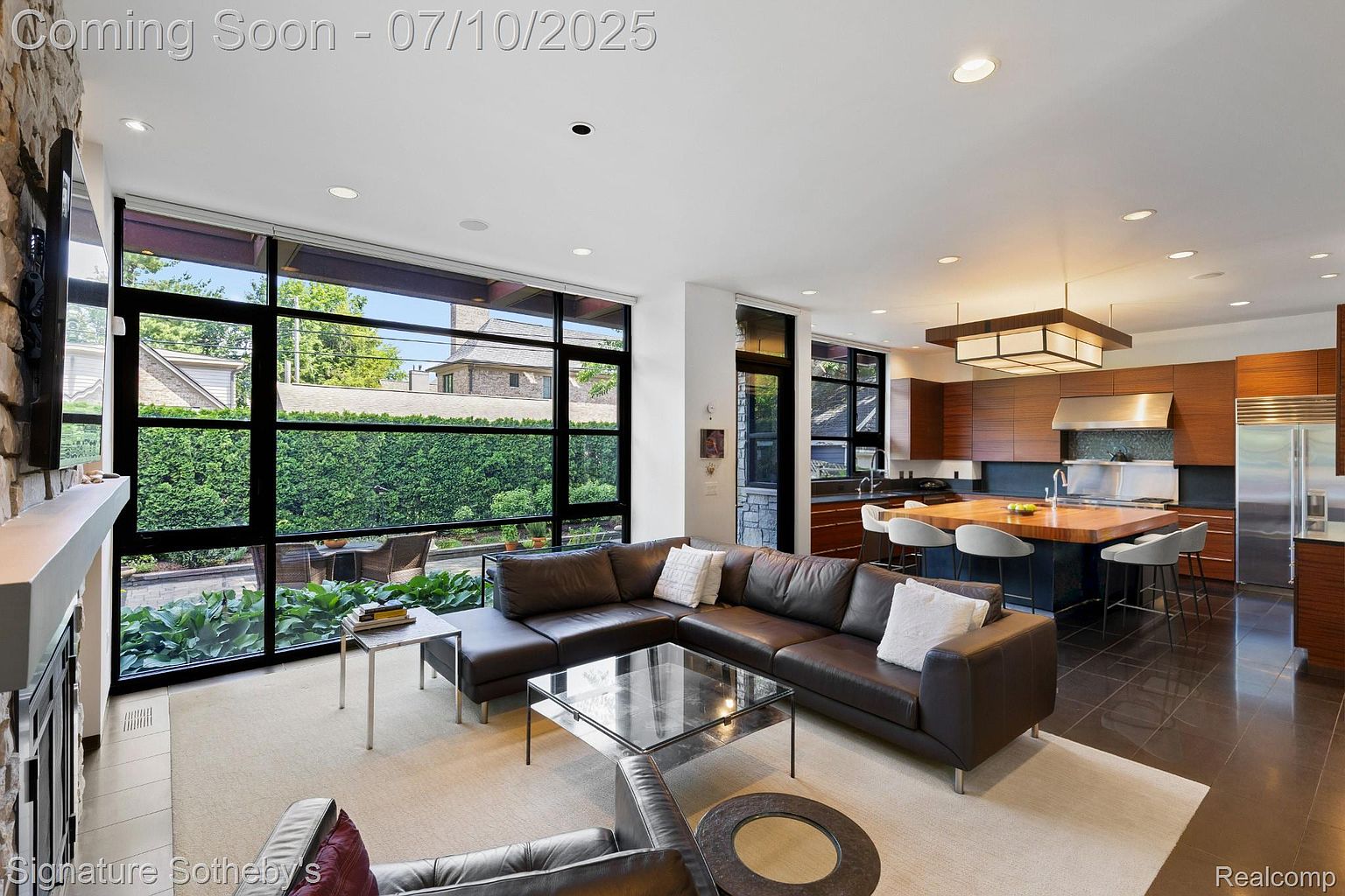 This is an interior shot of a modern living room that seamlessly blends with an open-concept kitchen. The living room features a large, dark leather sectional sofa, a glass coffee table, and expansive windows offering views of the outdoor greenery. The kitchen boasts sleek wooden cabinetry, a large island with bar seating, and stainless steel appliances, creating a sophisticated and inviting atmosphere.