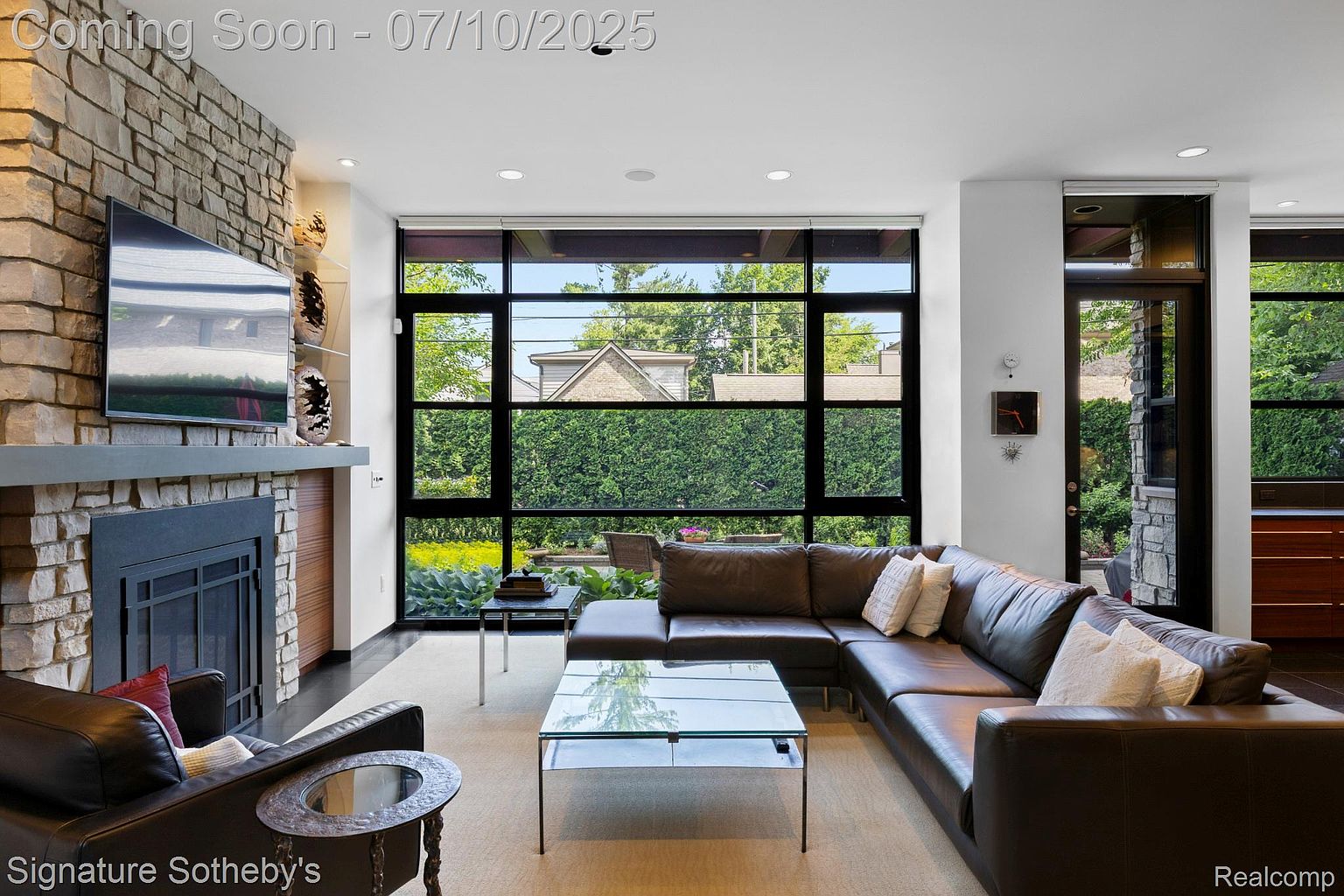This is an interior shot of a living room featuring a large sectional sofa, a glass coffee table, and a stone fireplace with a mounted television. A large window provides ample natural light and a view of the exterior greenery. The room exudes a modern and comfortable atmosphere.