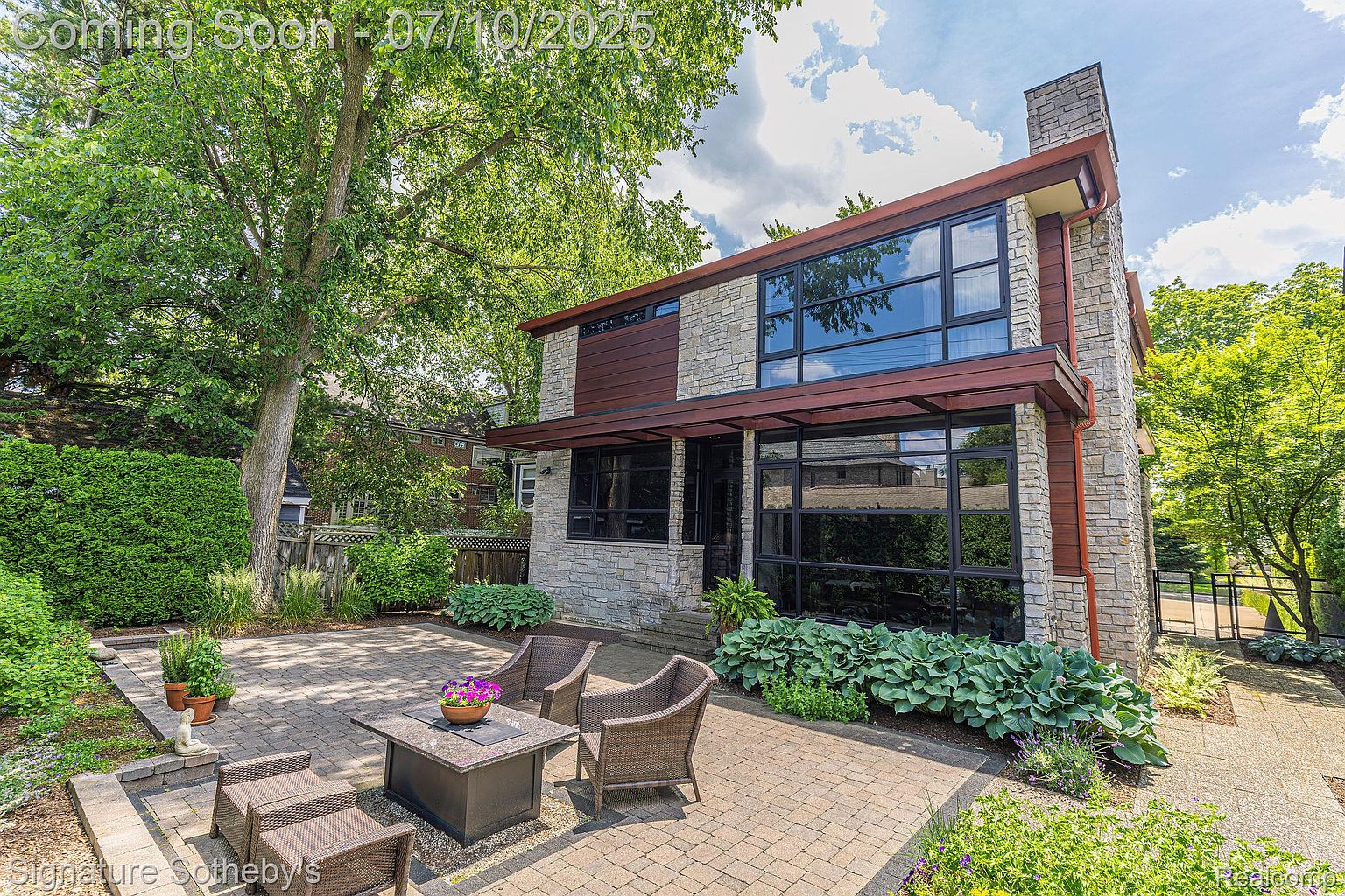 This image showcases a beautifully designed patio area adjacent to a modern home. The patio features brick pavers, comfortable outdoor seating arranged around a fire pit, and lush landscaping that enhances the outdoor living space. The home's exterior is characterized by a combination of stone and wood, with large windows that provide ample natural light.