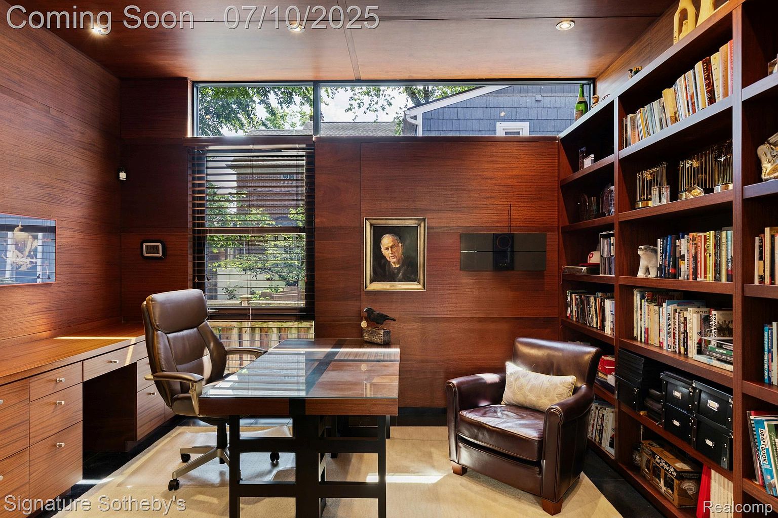 This is an interior shot of a sophisticated home office featuring rich wood paneling and built-in bookshelves filled with books and decorative items. A glass-topped desk with a leather chair sits in the center, complemented by a comfortable leather armchair. The room is well-lit with natural light streaming through a large window, creating a warm and inviting workspace.