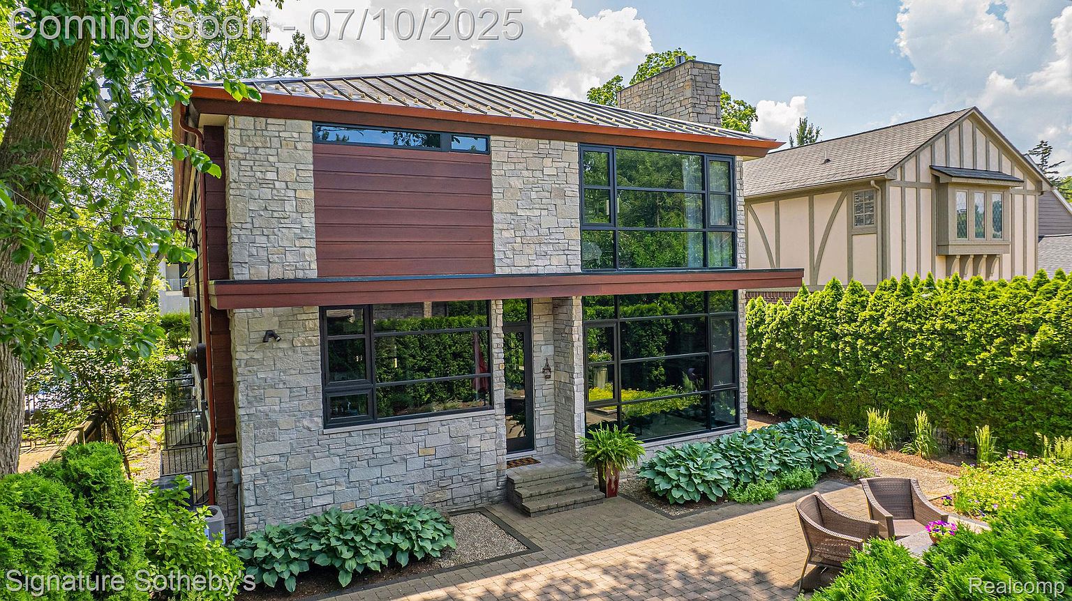 This is a front exterior view of a modern two-story home with a stone and wood facade. Large windows provide ample natural light, and the landscaping is well-maintained with lush greenery. A paved walkway leads to the front entrance, enhancing the property's curb appeal.