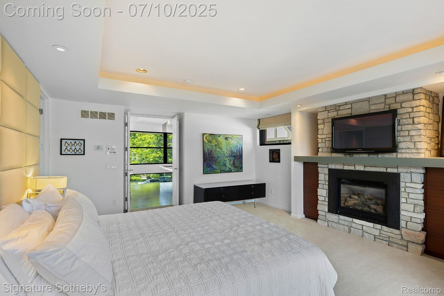 This is a primary bedroom featuring a king-size bed with white linens, a stone fireplace with a mounted television, and a door leading to an outdoor area. The room has a modern design with recessed lighting, a neutral color palette, and artwork on the walls. The overall impression is luxurious and comfortable.