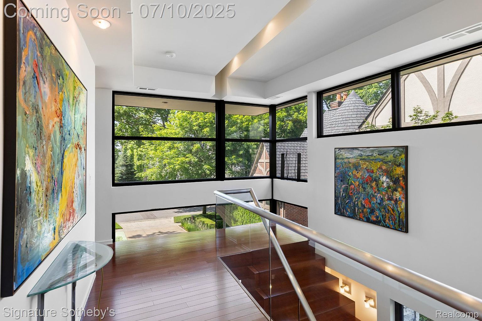 This interior shot showcases a modern hallway and staircase area with hardwood flooring and a glass railing. Large windows provide ample natural light and views of the surrounding greenery. Artwork adorns the walls, adding a touch of sophistication to the space.