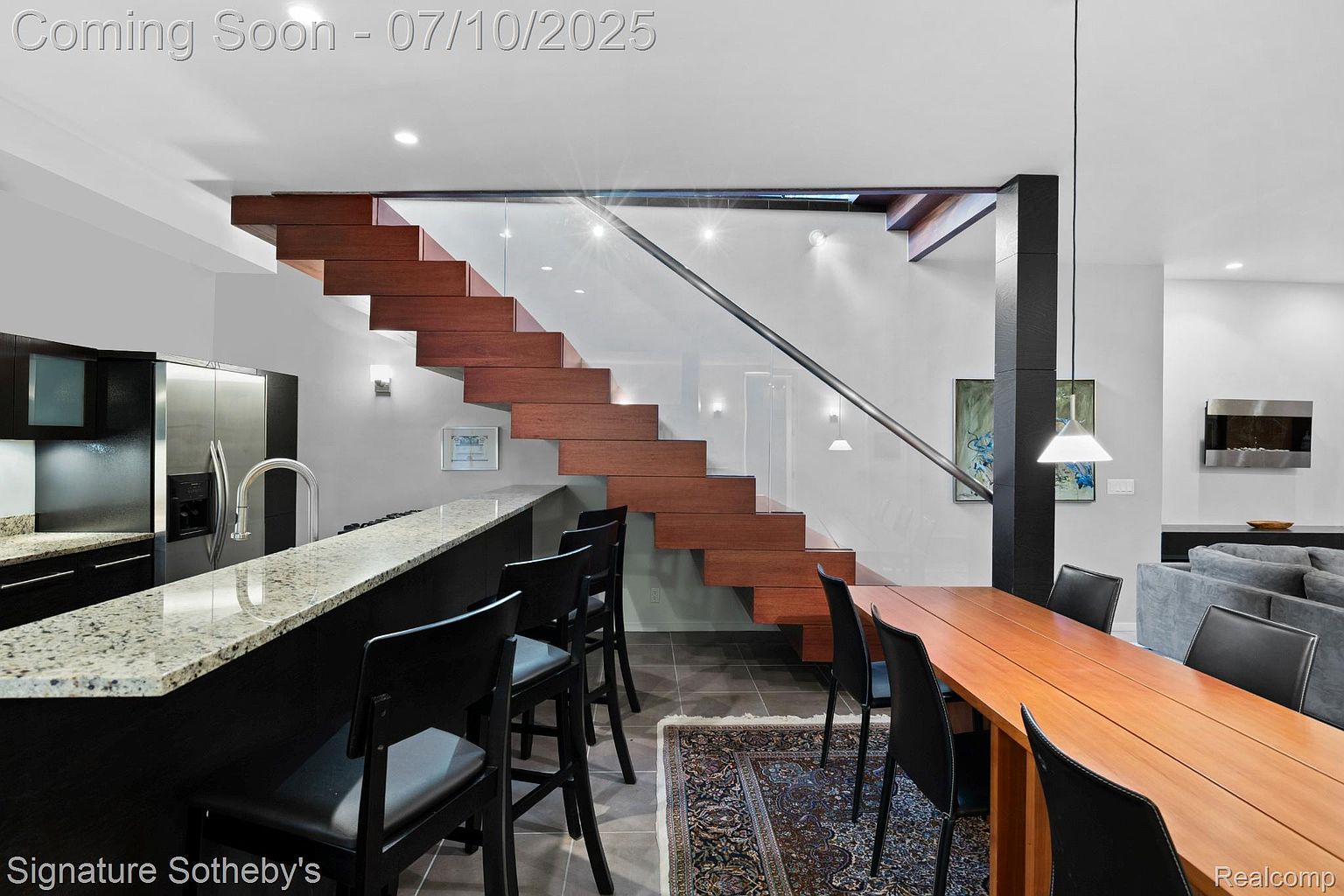This interior shot showcases a modern dining area with a unique floating staircase as a focal point. The dining table is made of wood and is surrounded by black chairs, complemented by a stylish kitchen bar with granite countertops and bar stools. The room features a contemporary design with a blend of textures and materials, creating an inviting and sophisticated atmosphere.