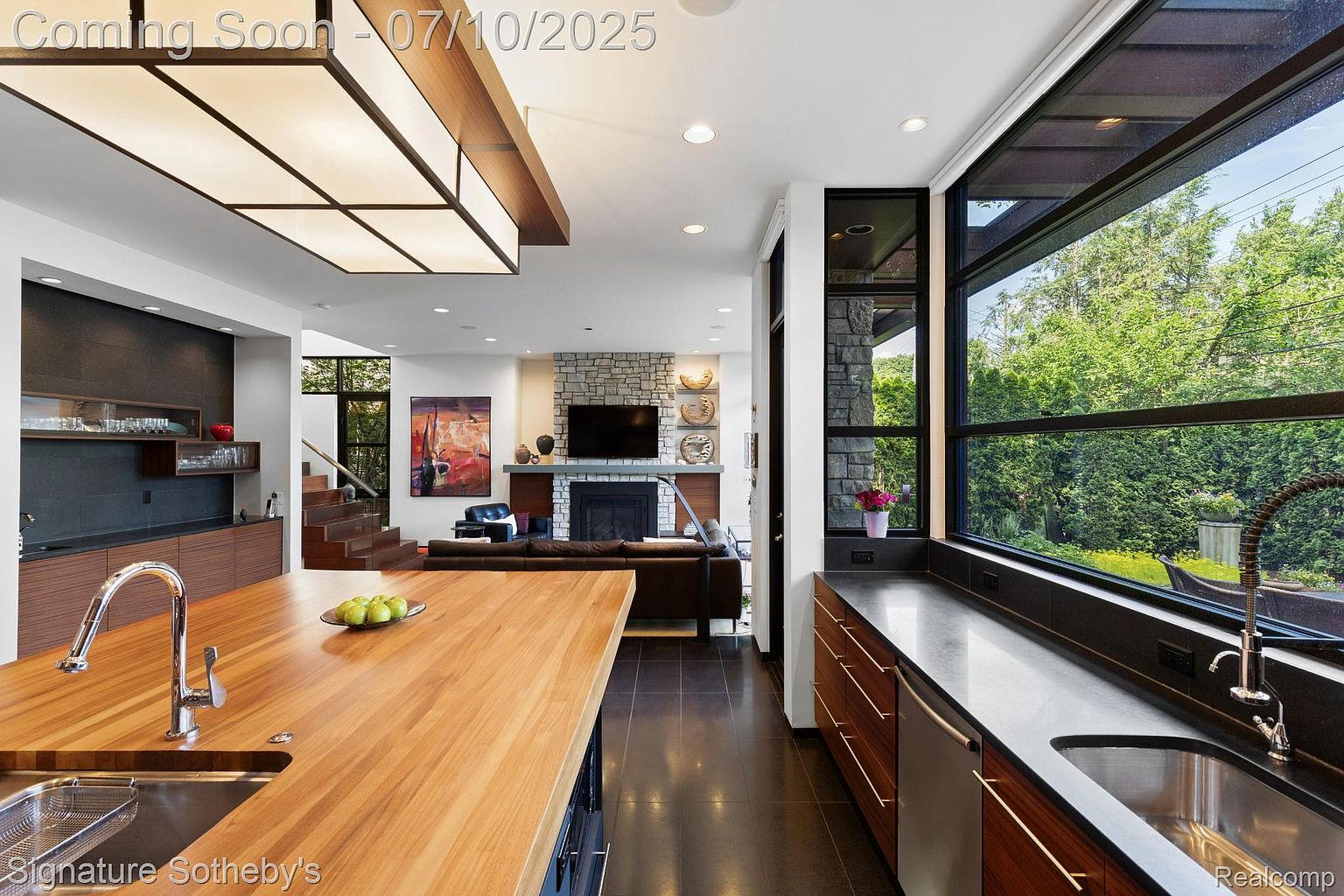 This interior shot showcases a modern kitchen that seamlessly transitions into a living area. The kitchen features a large wooden island with a sink, sleek cabinetry, and a window offering a view of the outdoors. The living area includes a stone fireplace, a television, and comfortable seating, creating an open and inviting space.