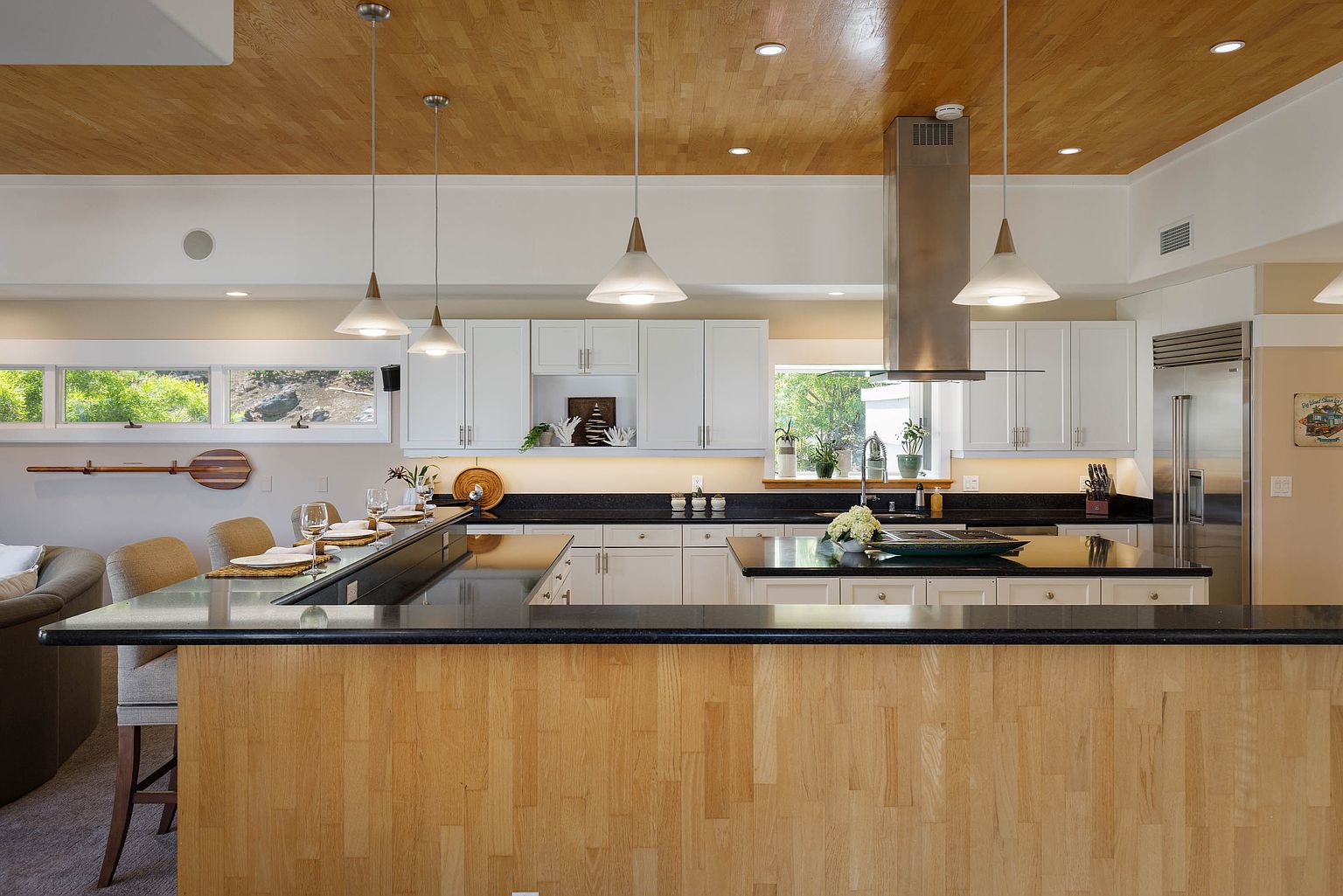 This is a well-lit kitchen featuring a wooden ceiling and white cabinetry. A large island with a black countertop and wooden base provides ample workspace and seating. Stainless steel appliances and pendant lighting add a modern touch to the space, while windows offer natural light and views of the outdoors.