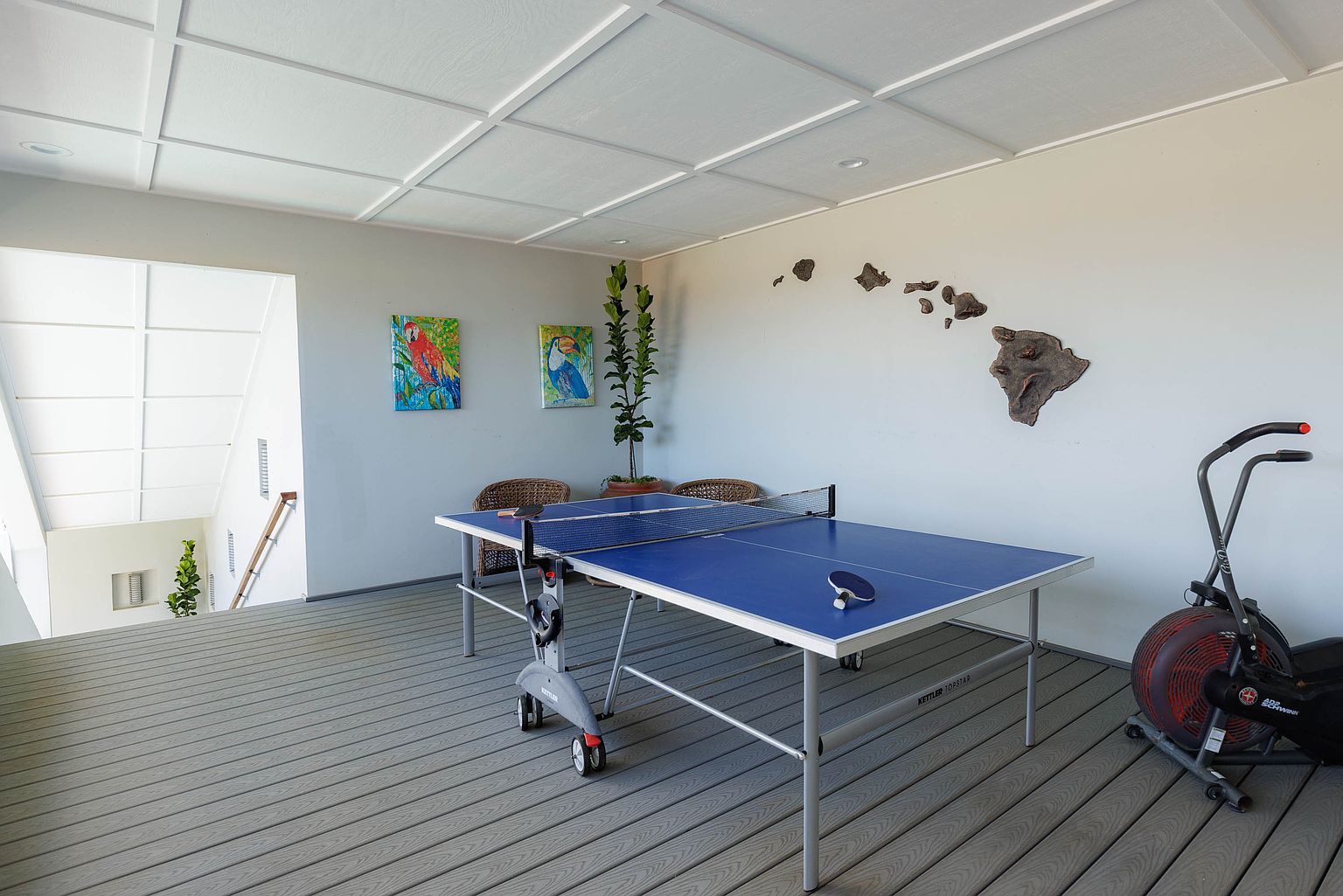 This interior shot showcases a home gym area featuring a ping pong table and an exercise bike. The room is well-lit with a modern ceiling design and decorated with artwork and a wooden map of Hawaii. The flooring is a gray wood-look material, creating a clean and inviting space for recreation and fitness.