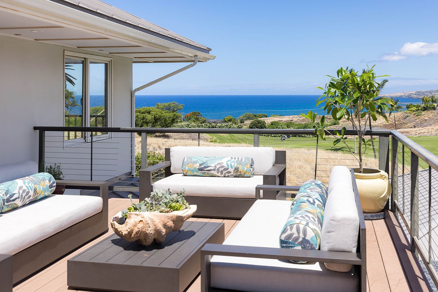 This image showcases a luxurious outdoor patio or deck area with comfortable seating arrangements, including sofas and chairs with stylish cushions. The space overlooks a stunning ocean view and a well-maintained golf course, creating a serene and upscale ambiance. The deck is furnished with a coffee table featuring a unique shell planter, and a potted tree adds a touch of greenery.