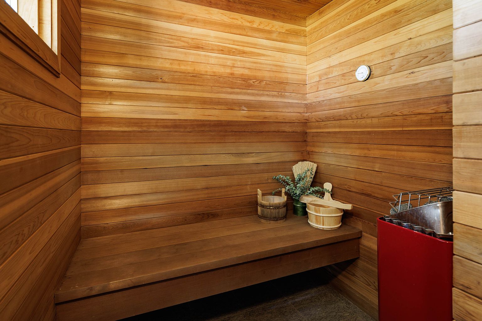 This is an interior shot of a sauna, featuring wooden walls and a built-in wooden bench. A red sauna heater is visible in the corner, along with sauna accessories like buckets and a whisk. The space is well-lit, creating a warm and inviting atmosphere, perfect for relaxation and wellness.