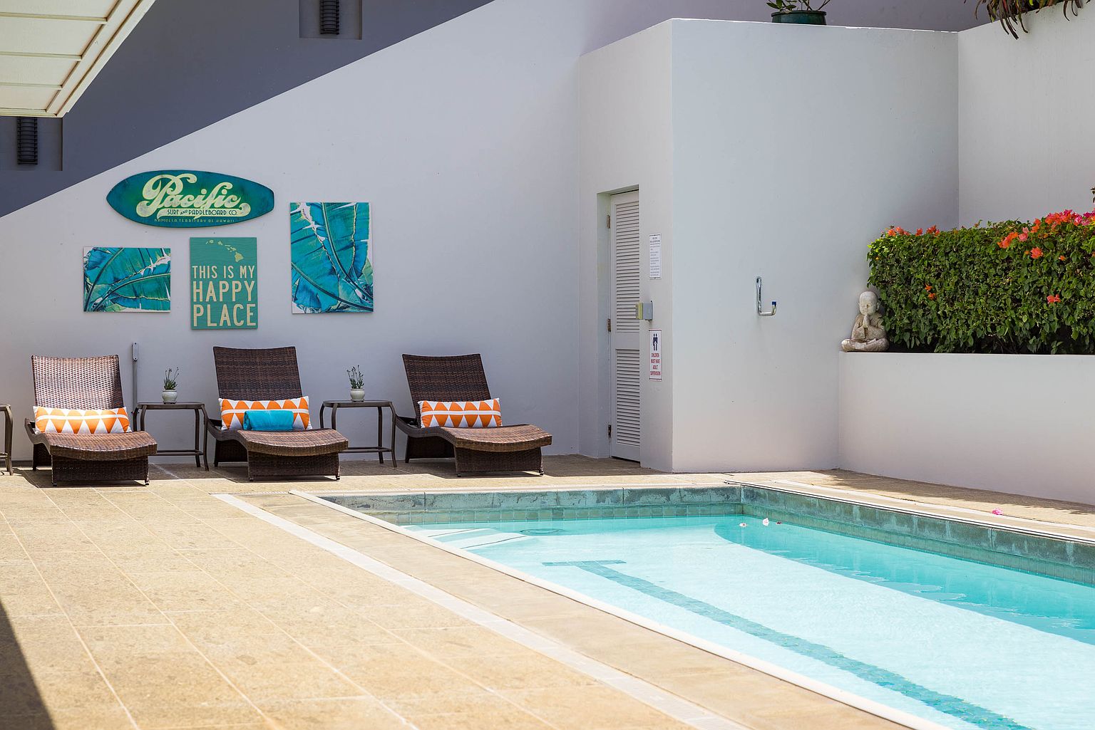 This image showcases a serene outdoor pool area, featuring three wicker lounge chairs with decorative pillows, small side tables, and tropical-themed artwork adorning the wall. The pool's clear blue water invites relaxation, while a small Buddha statue and lush greenery add a touch of tranquility. The perspective is a medium shot, capturing the entire pool area and its surrounding elements.