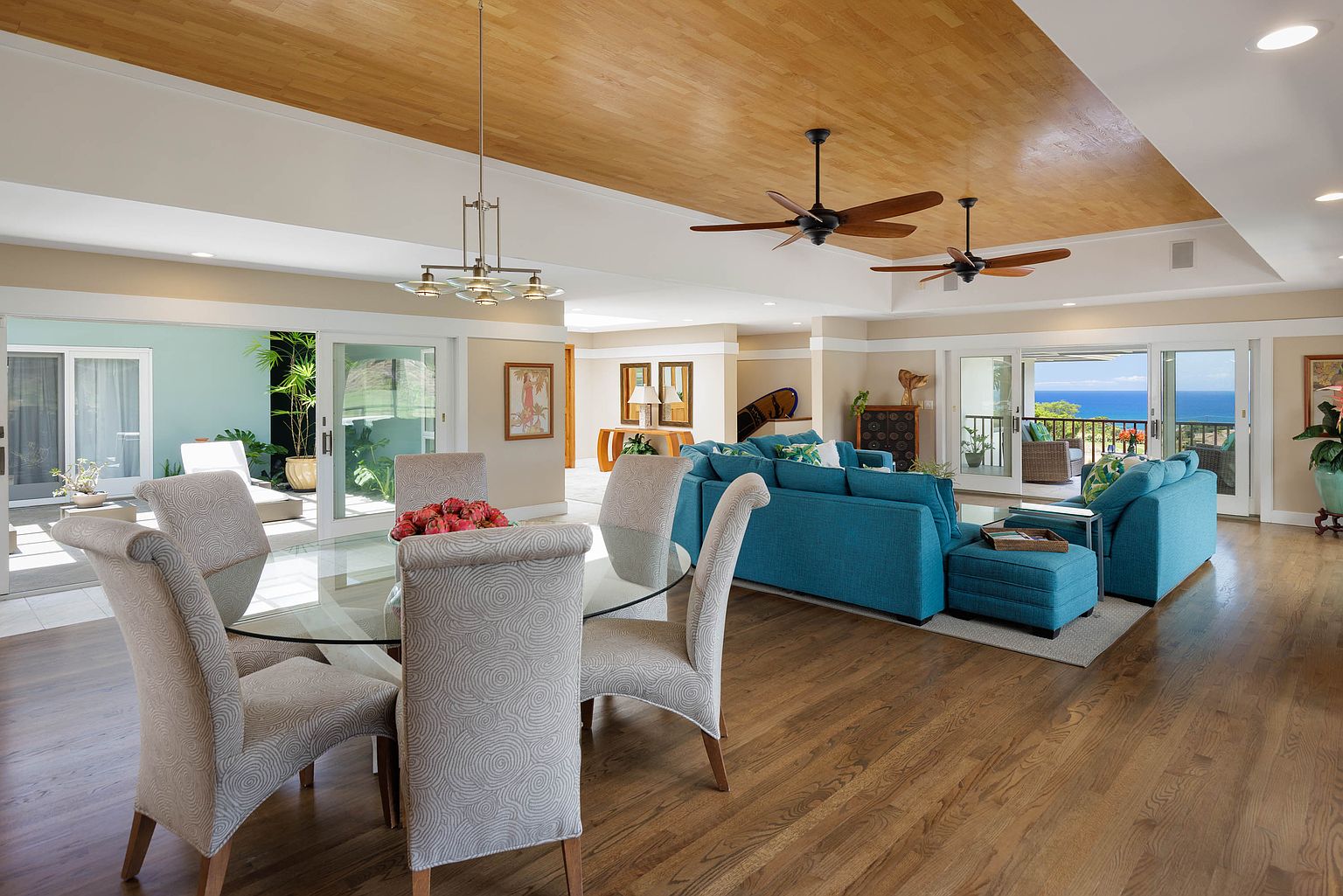 This is a spacious and bright living room featuring hardwood floors, a wooden ceiling, and large sliding glass doors that offer an ocean view. The room is furnished with a blue sectional sofa, a glass-top dining table with upholstered chairs, and ceiling fans. The open layout and natural light create an inviting and luxurious atmosphere.