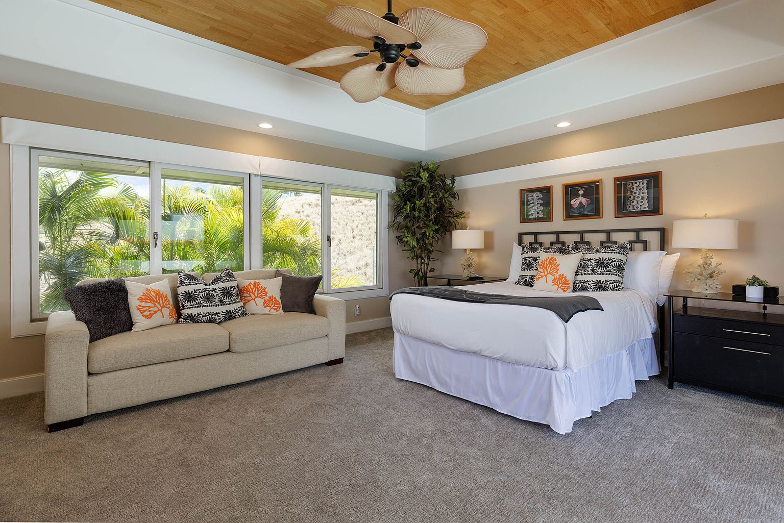 This primary bedroom features a neutral color palette with a beige sofa, a bed with white linens and decorative pillows, and dark wood furniture. Large windows provide ample natural light and views of lush greenery. The room is spacious and well-appointed, creating a relaxing and inviting atmosphere.