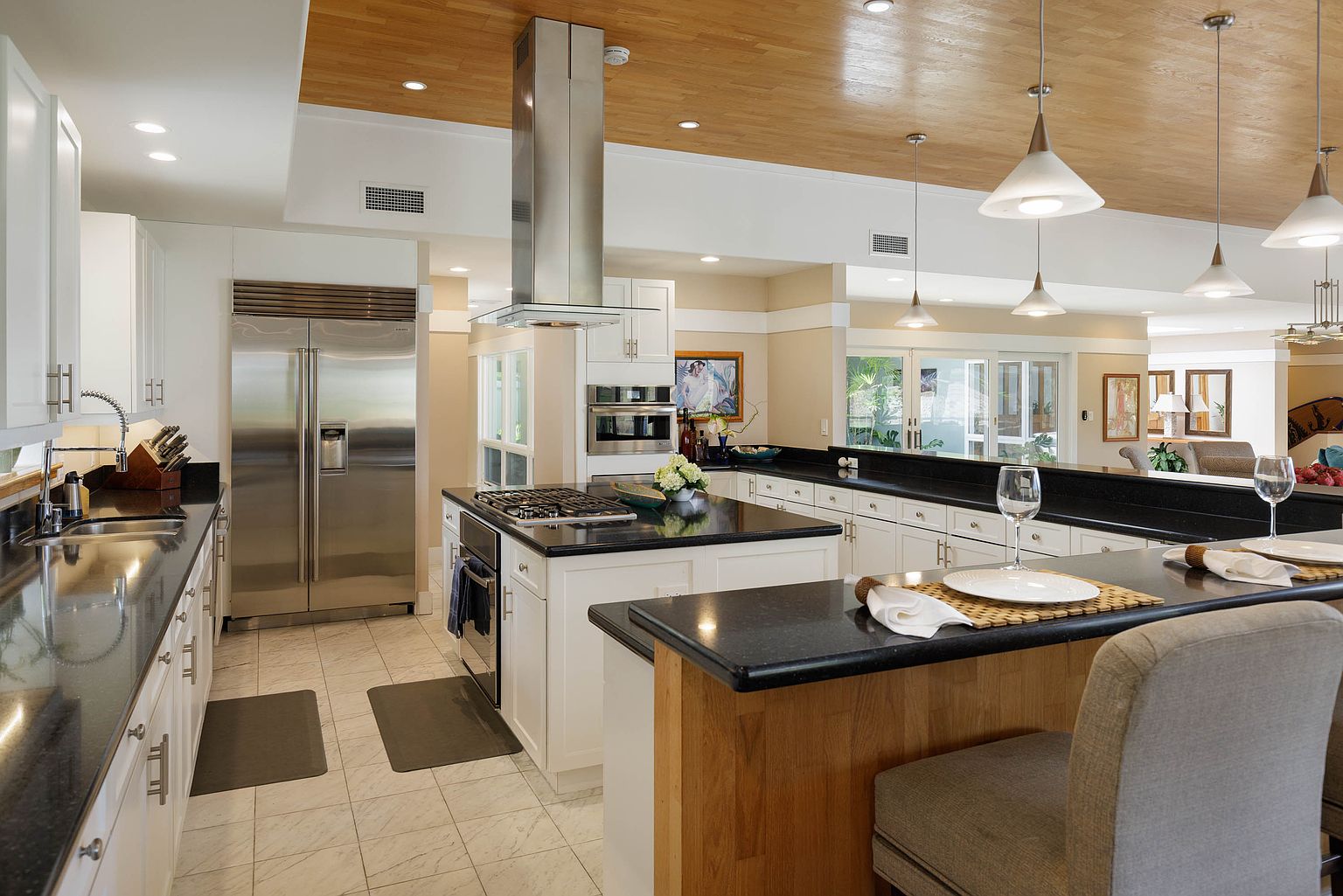 This is a well-lit kitchen featuring white cabinetry, stainless steel appliances, and a dark countertop. A kitchen island with a cooktop and a breakfast bar provides ample workspace and seating. The wooden ceiling and pendant lights add a touch of warmth to the modern design.
