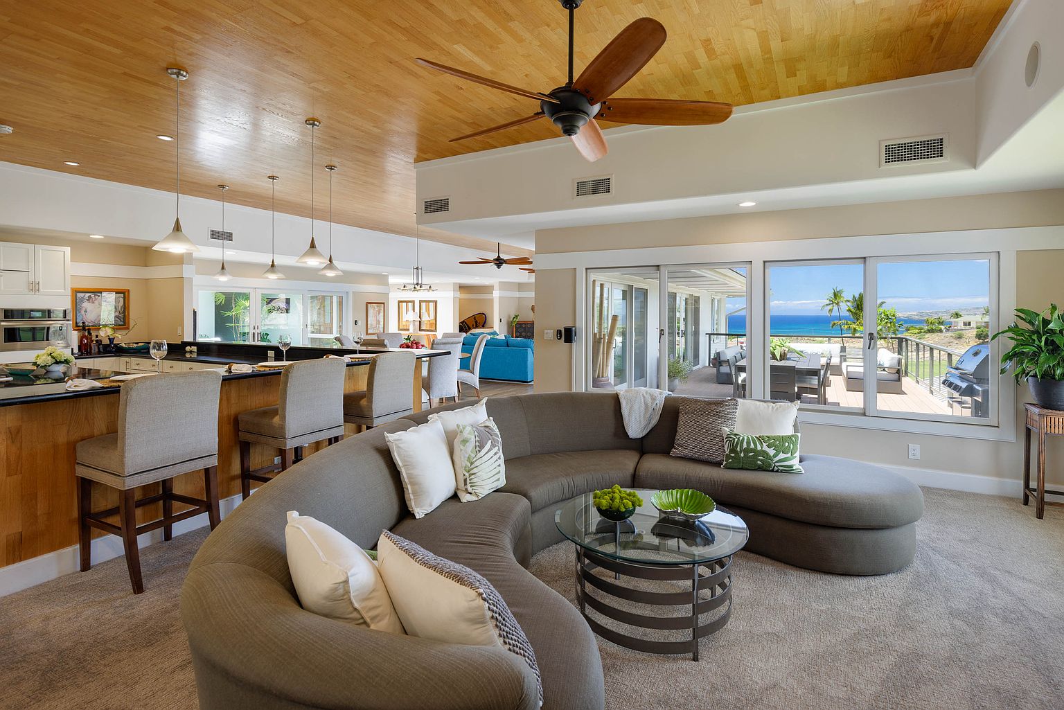 This is an interior shot of a luxurious living room featuring a curved sectional sofa, a glass-topped coffee table, and a view of the ocean through large windows. The room has a warm and inviting atmosphere, with a wooden ceiling and neutral color palette, creating a seamless transition to the outdoor deck area.