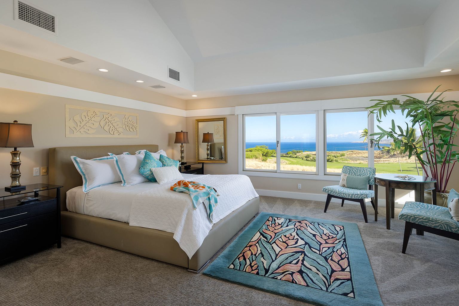 This is a bright and airy primary bedroom featuring a king-size bed with white linens and turquoise accents. The room has a neutral color palette with beige walls and carpet, complemented by a large window offering stunning ocean and golf course views. The room is tastefully decorated with lamps, a mirror, and a rug with a floral pattern, creating a serene and luxurious atmosphere.