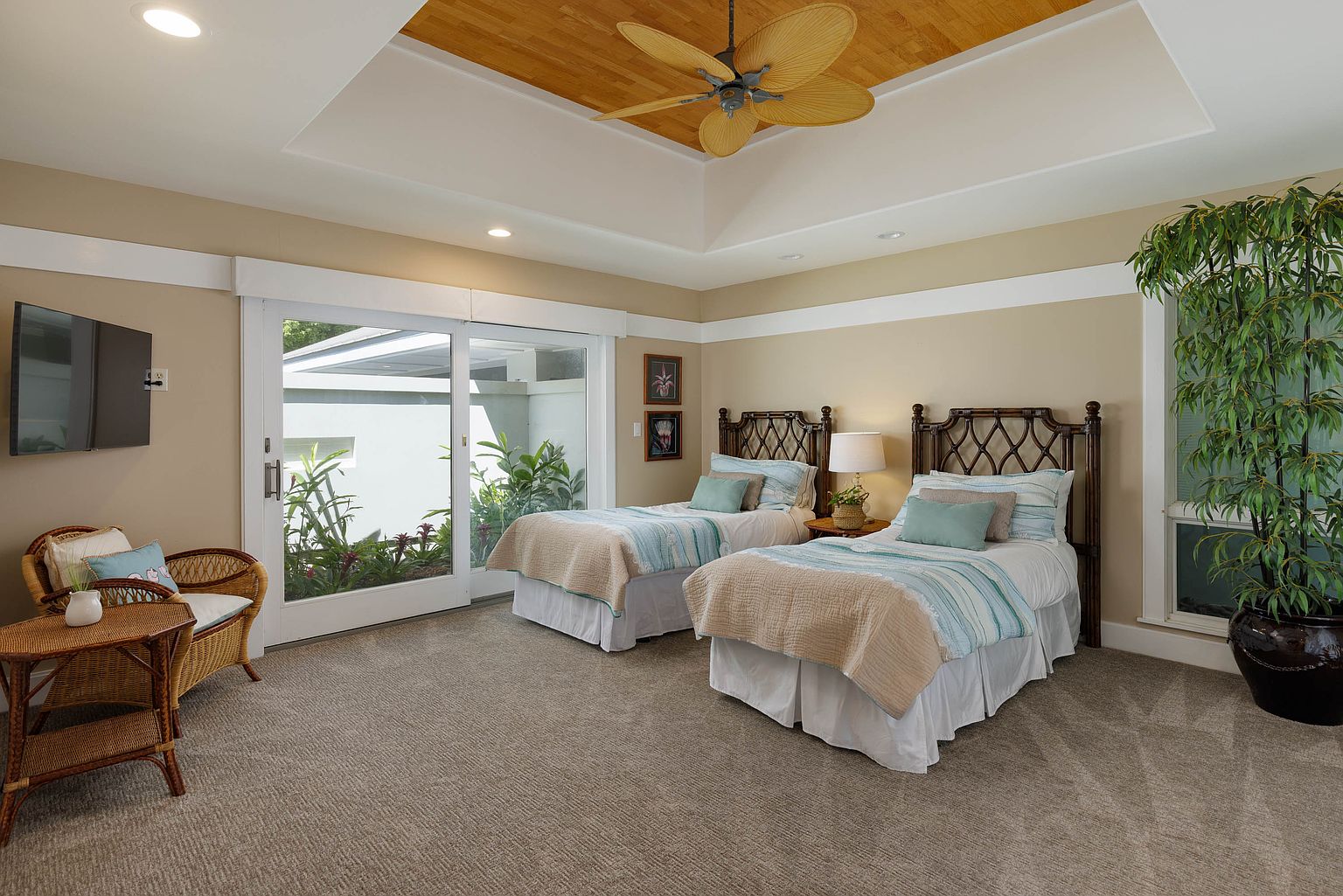 This is a cozy guest bedroom featuring two twin beds with decorative headboards, light blue bedding, and neutral-toned throws. A sliding glass door provides natural light and a view of the outdoor greenery. The room is decorated in a simple, comfortable style with a ceiling fan and a warm color palette, creating a welcoming atmosphere.