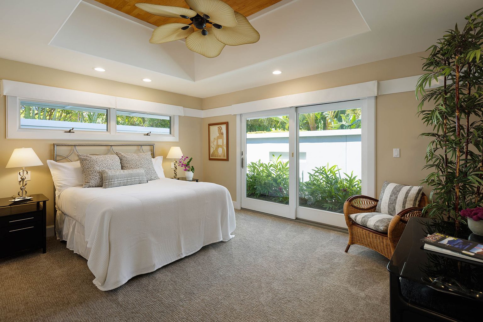 This is a well-lit primary bedroom featuring a king-size bed with a white comforter and decorative pillows. The room has a neutral color palette with beige walls and carpet, complemented by white trim and a unique ceiling fan. Sliding glass doors provide access to the outside, enhancing the room's bright and airy feel.