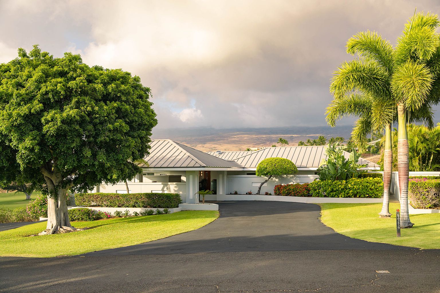 This image showcases the front exterior of a modern, single-story home with a well-manicured lawn and mature landscaping. A curved driveway leads to the entrance, flanked by lush greenery and palm trees. The home features a light-colored facade and a sleek, low-pitched roof, creating a serene and inviting curb appeal.