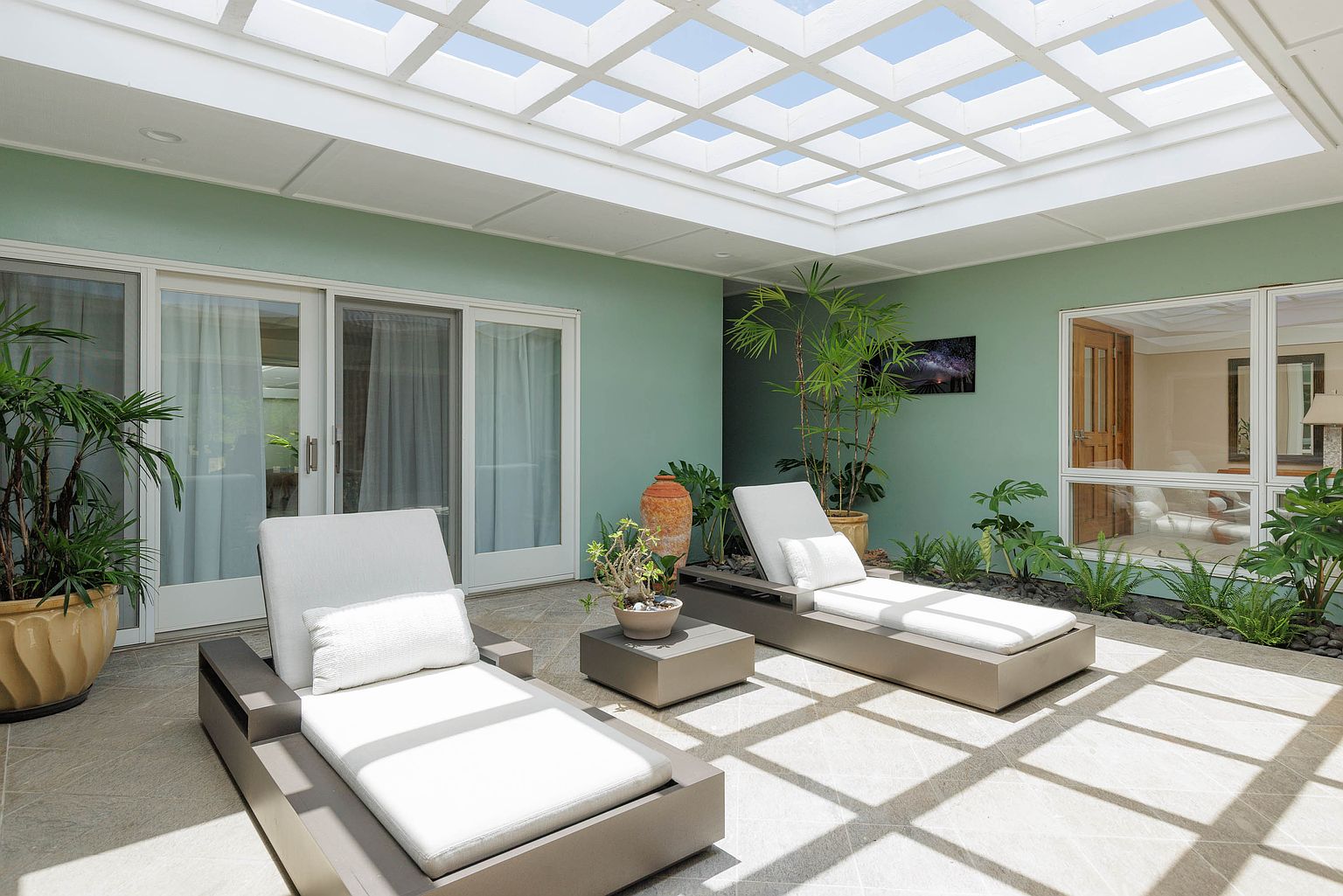 This image showcases a serene patio area featuring two modern lounge chairs with white cushions, arranged around a small side table. The space is enhanced by lush greenery and potted plants, creating a tranquil atmosphere. A unique grid-patterned skylight provides natural light, while sliding glass doors offer seamless indoor-outdoor living.
